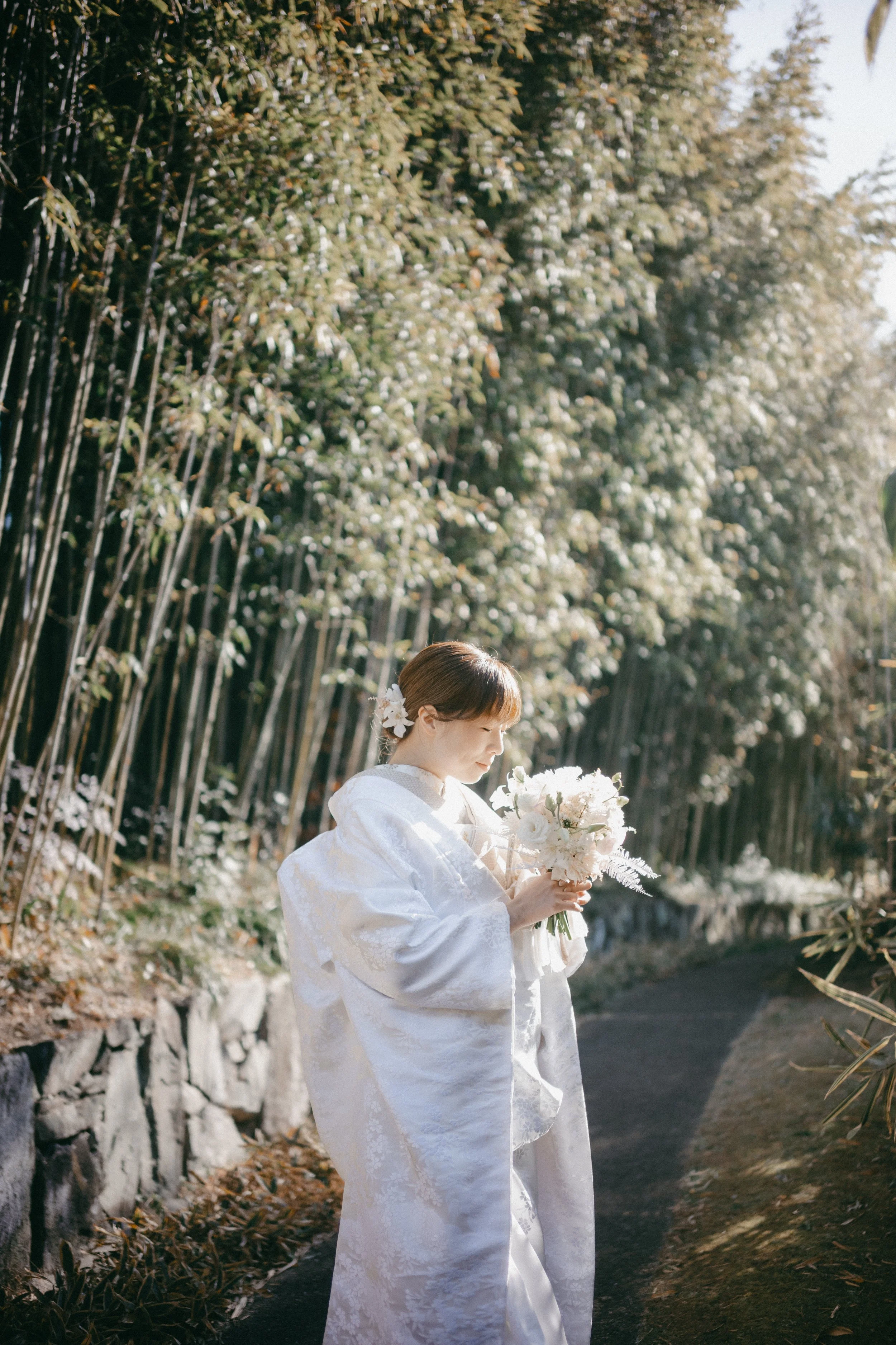 A woman dressed in a traditional white wedding kimono holding a bouquet of white flowers and posing outdoors along a path bordered by a bamboo fence and surrounded by lush greenery.