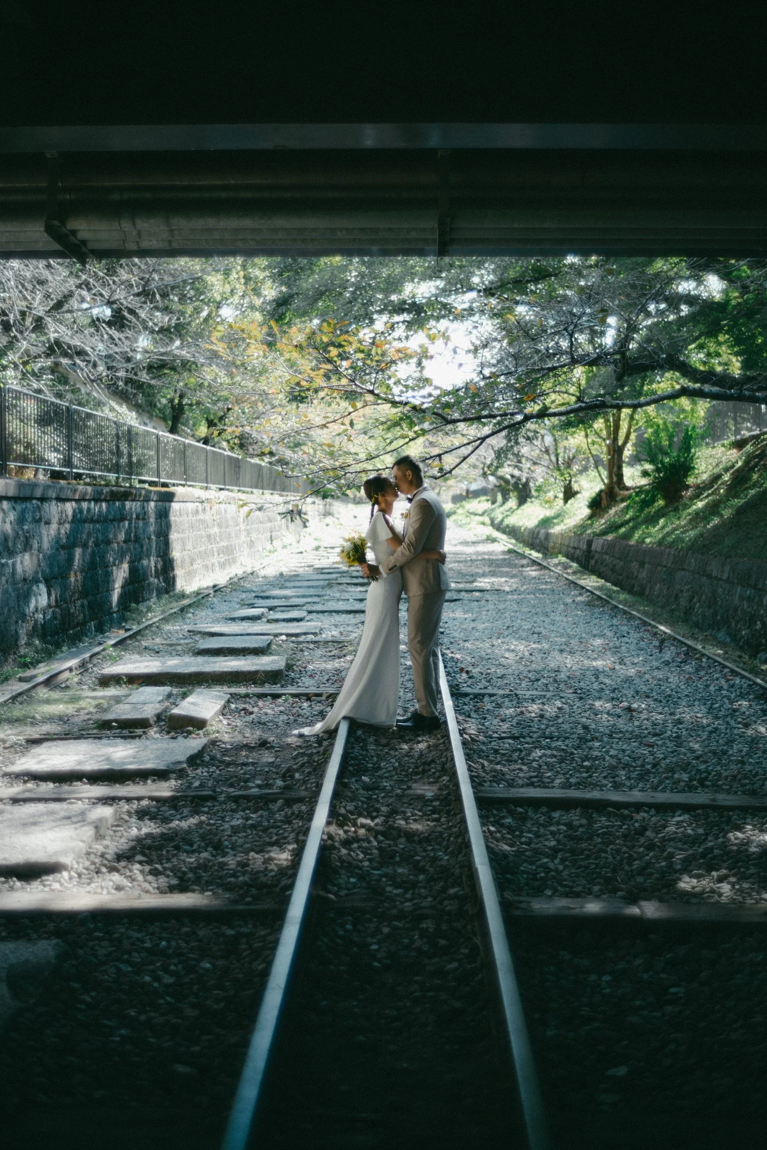A bride and groom standing on train tracks under a bridge, holding each other and smiling, with trees and sunlight in the background.
