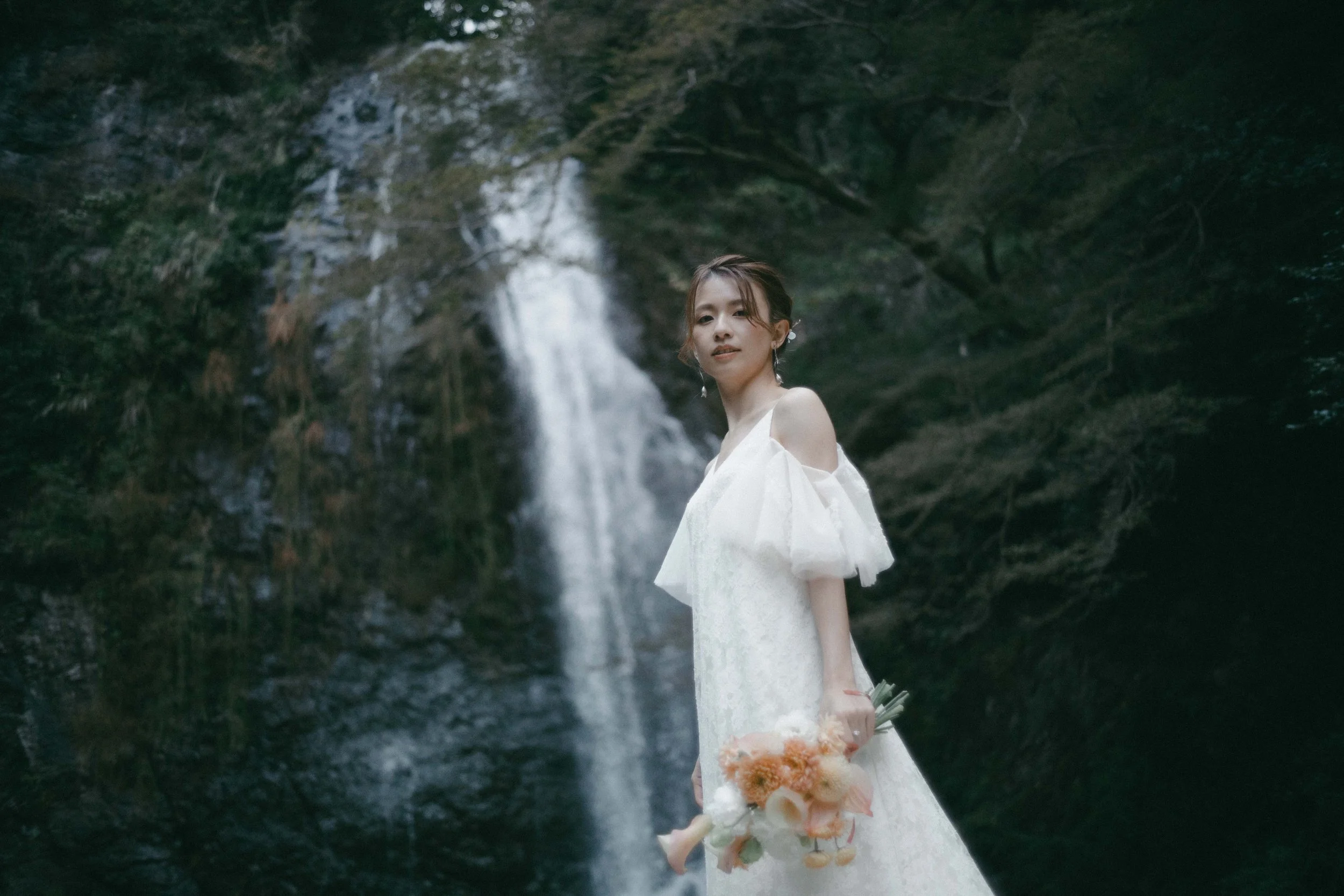 A woman in a white dress holding a bouquet standing in front of a waterfall surrounded by trees.