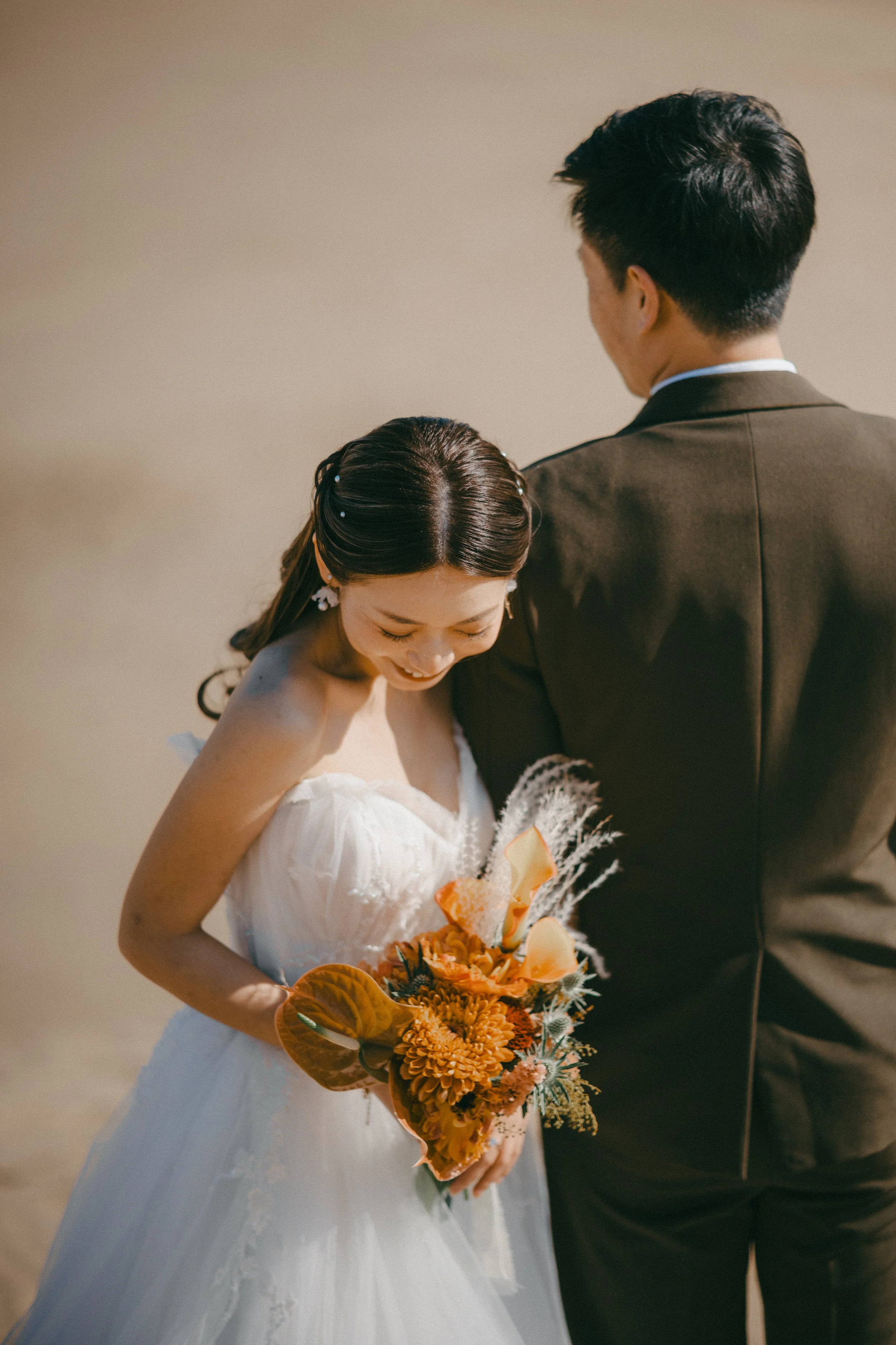 A bride in a white wedding dress holding a bouquet of orange and yellow flowers, standing next to a groom in a dark suit, both smiling with the bride looking down.
