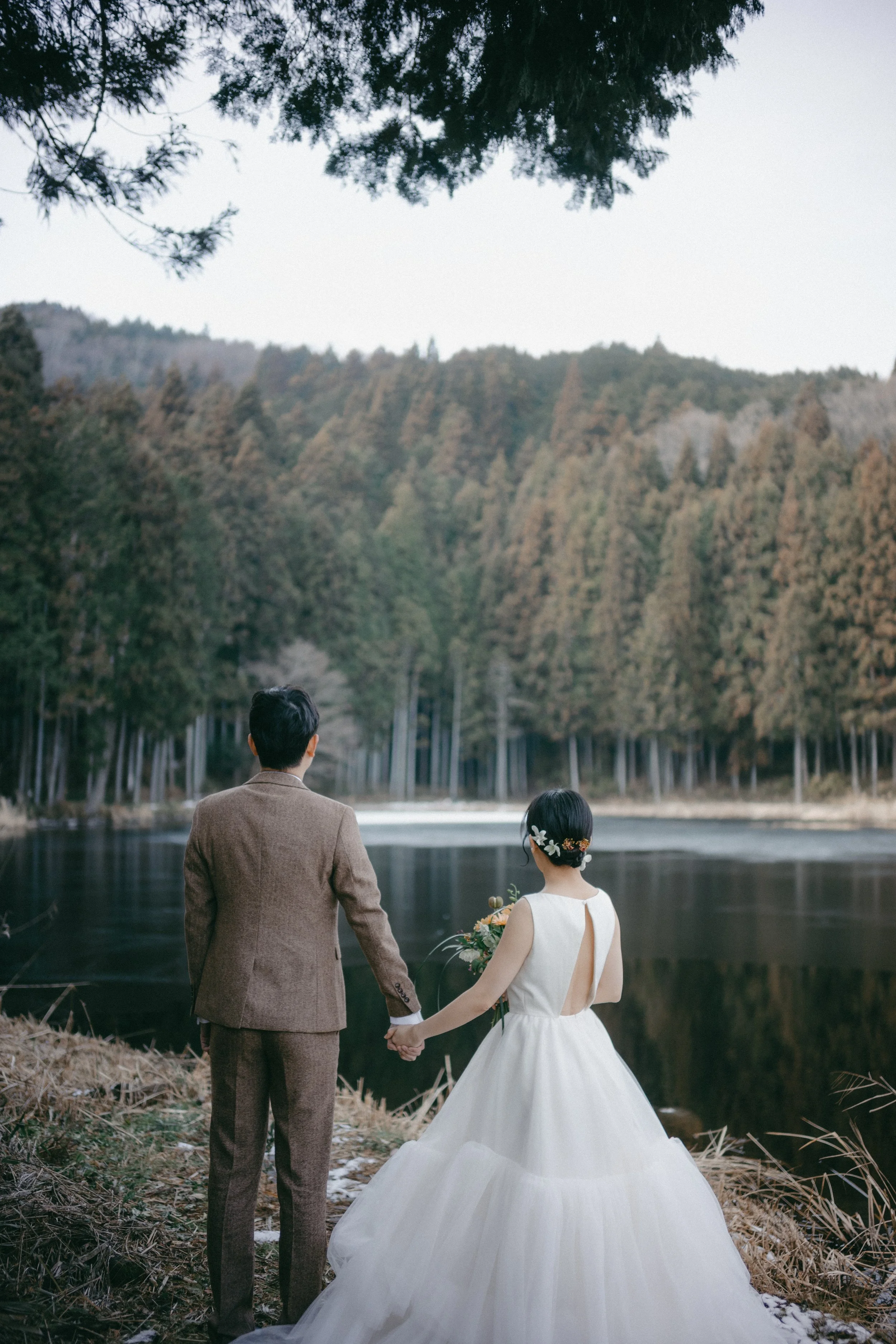A bride and groom standing by a lake, holding hands, with trees and mountains in the background.