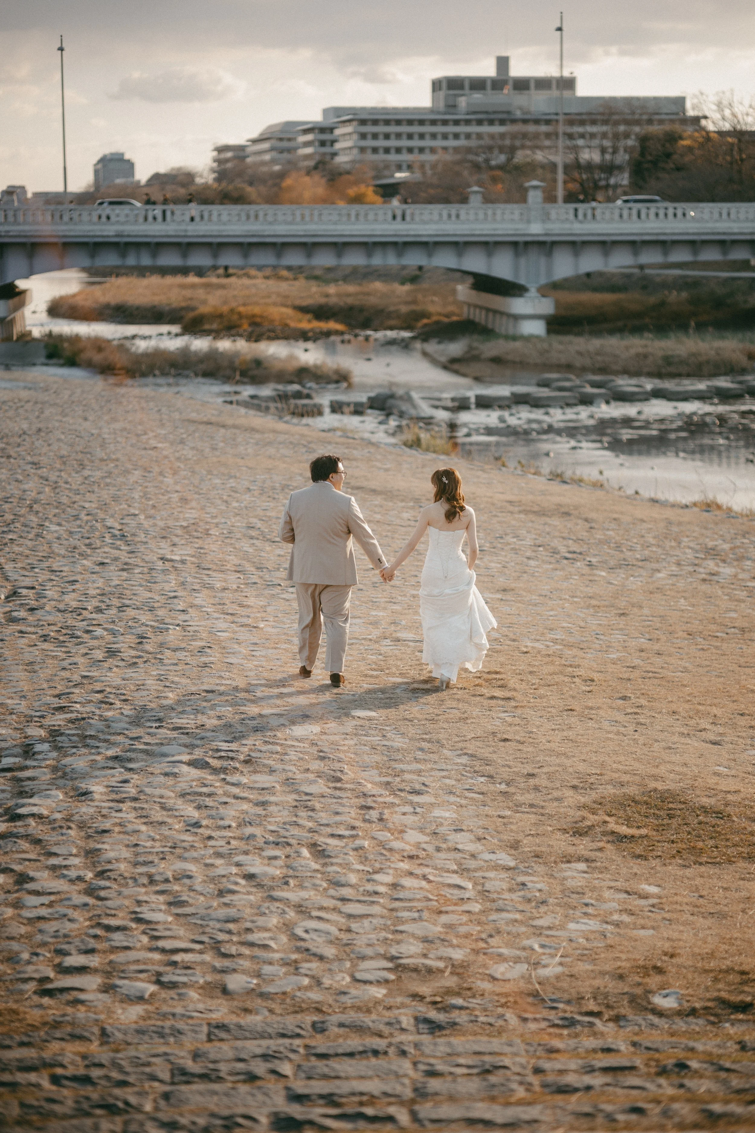 A couple in wedding attire holding hands while walking on a cobblestone path by a river