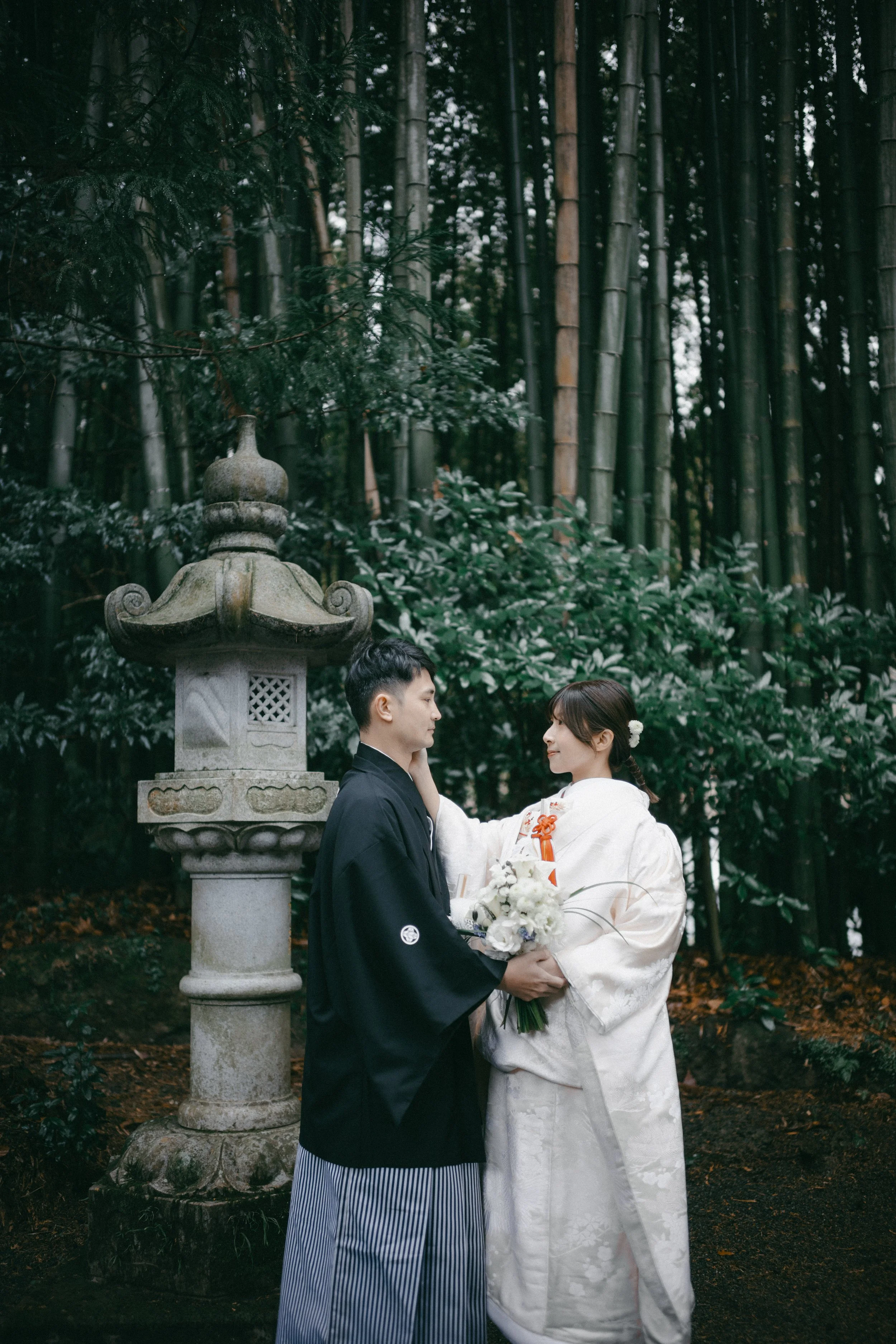 A couple dressed in traditional Japanese wedding attire standing in front of a stone lantern in a lush bamboo forest.
