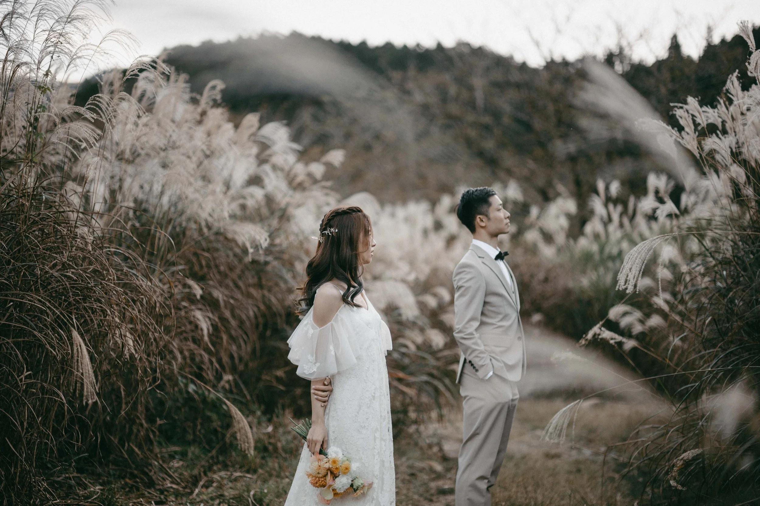 A bride and groom standing side by side in a neutral-colored outdoor wedding setting with tall grass and hills in the background, the bride holding a bouquet of flowers and wearing a white wedding dress, and the groom dressed in a light beige suit wi