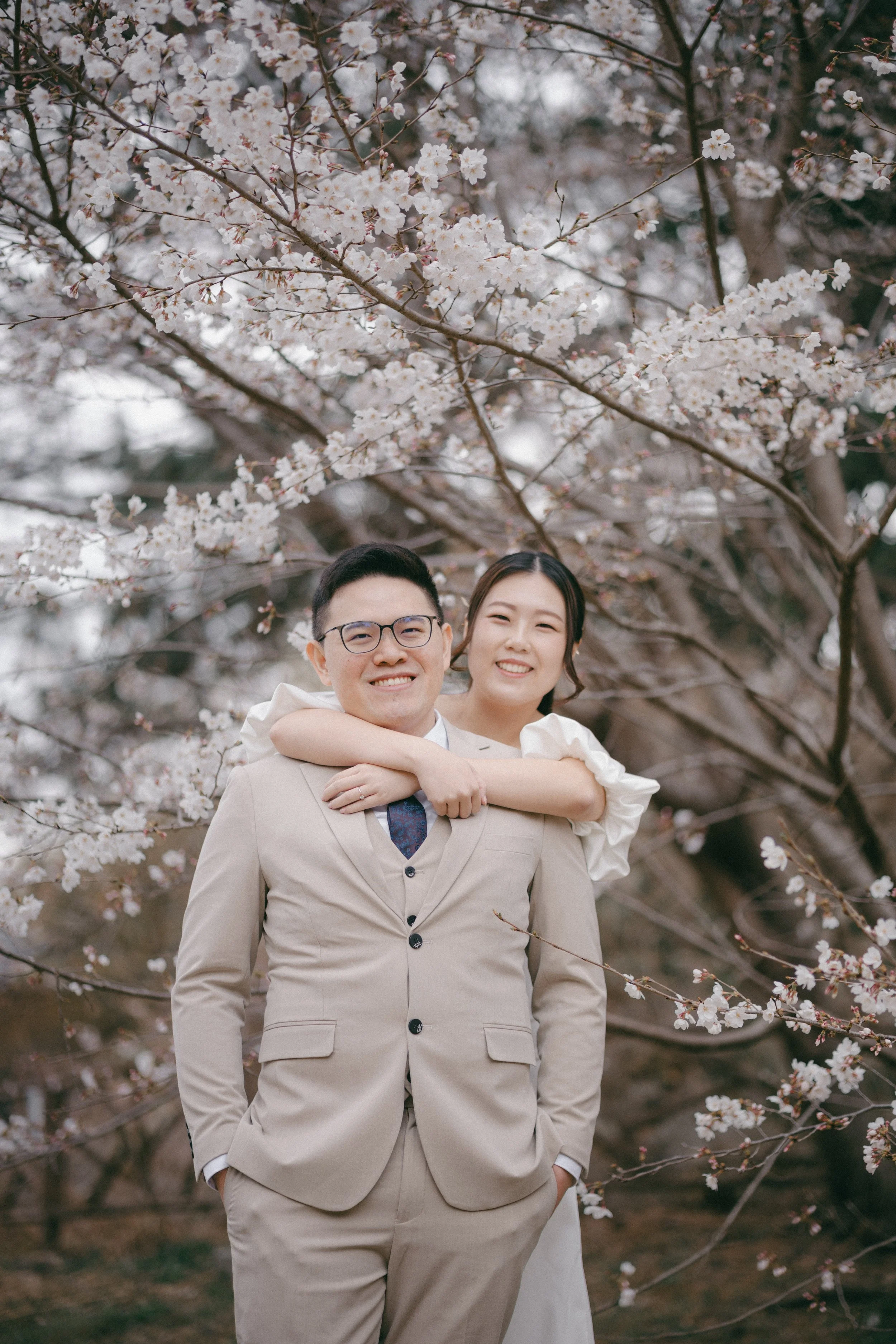 A smiling man in a beige suit with glasses, a woman in white with her arms around his neck, standing under blooming cherry blossom trees in spring.