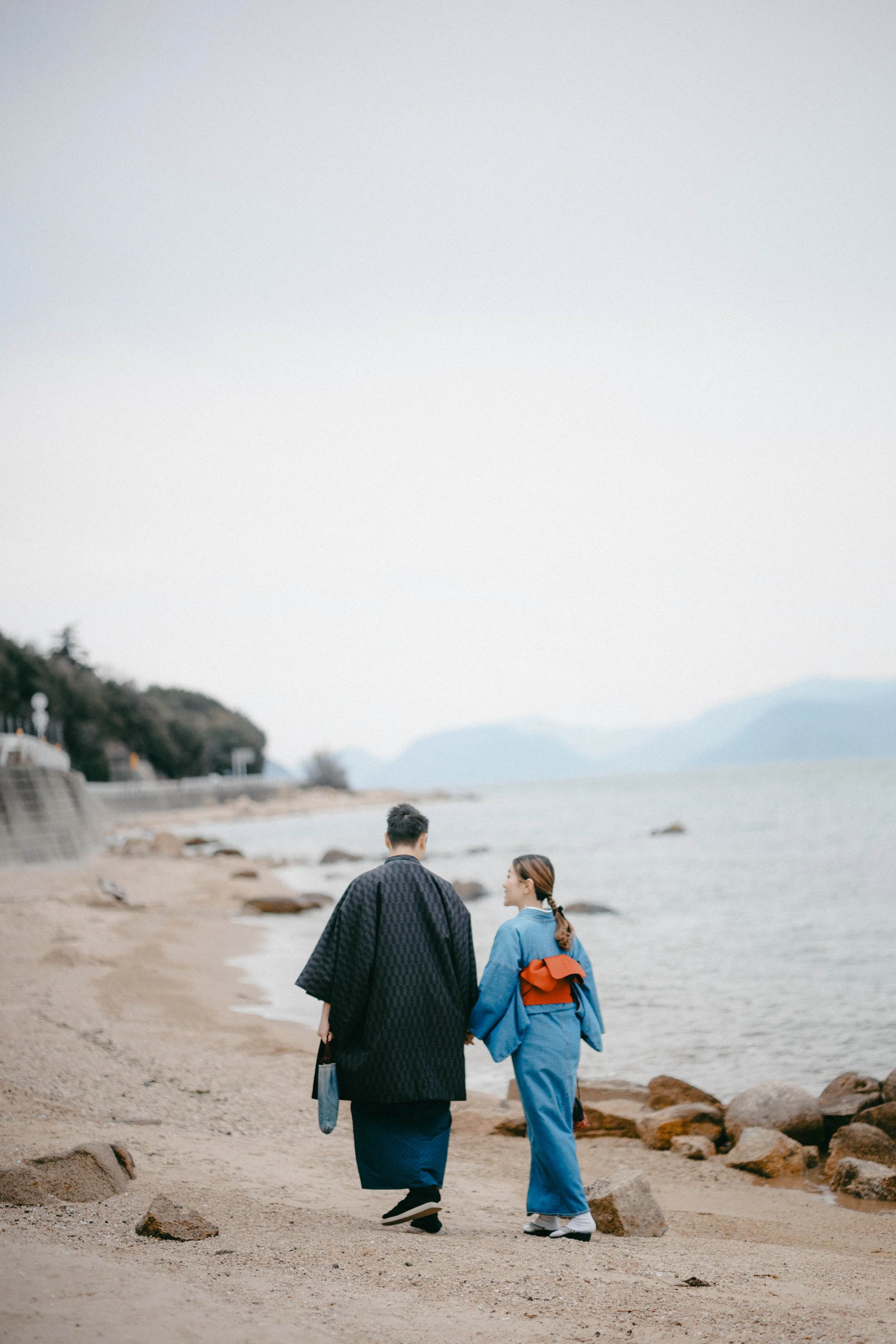 Two people walking on a sandy beach, wearing traditional Japanese kimonos, with the ocean and mountains in the background.