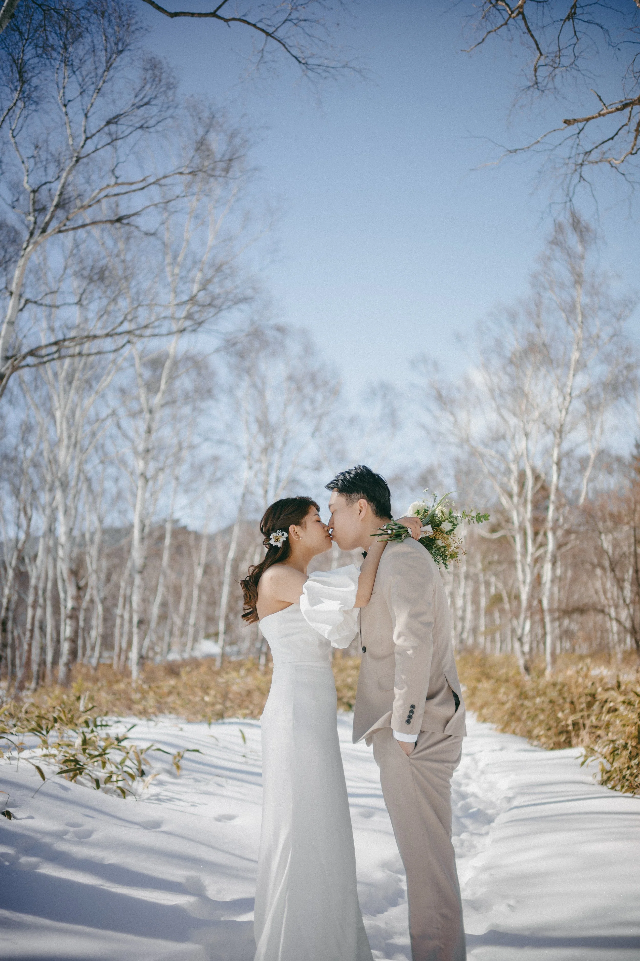 A bride and groom kissing in a snowy outdoor setting with leafless trees in the background.