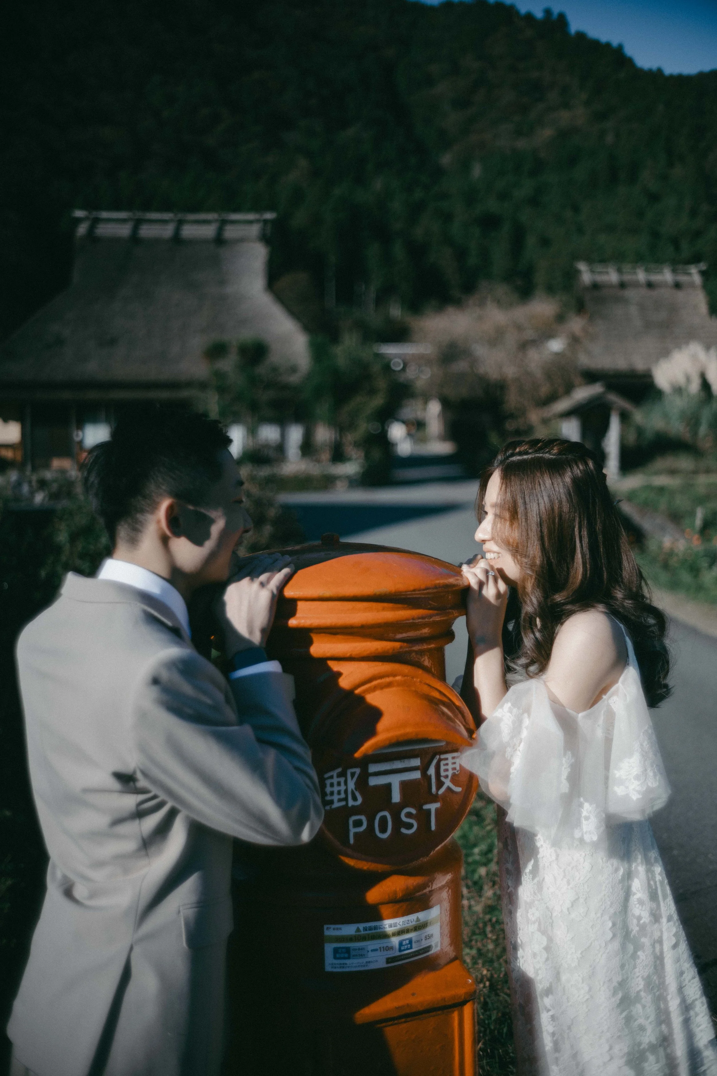 A young couple plays on a bright orange Japanese mailbox in a rural area with traditional thatched-roof houses and green mountains in the background.
