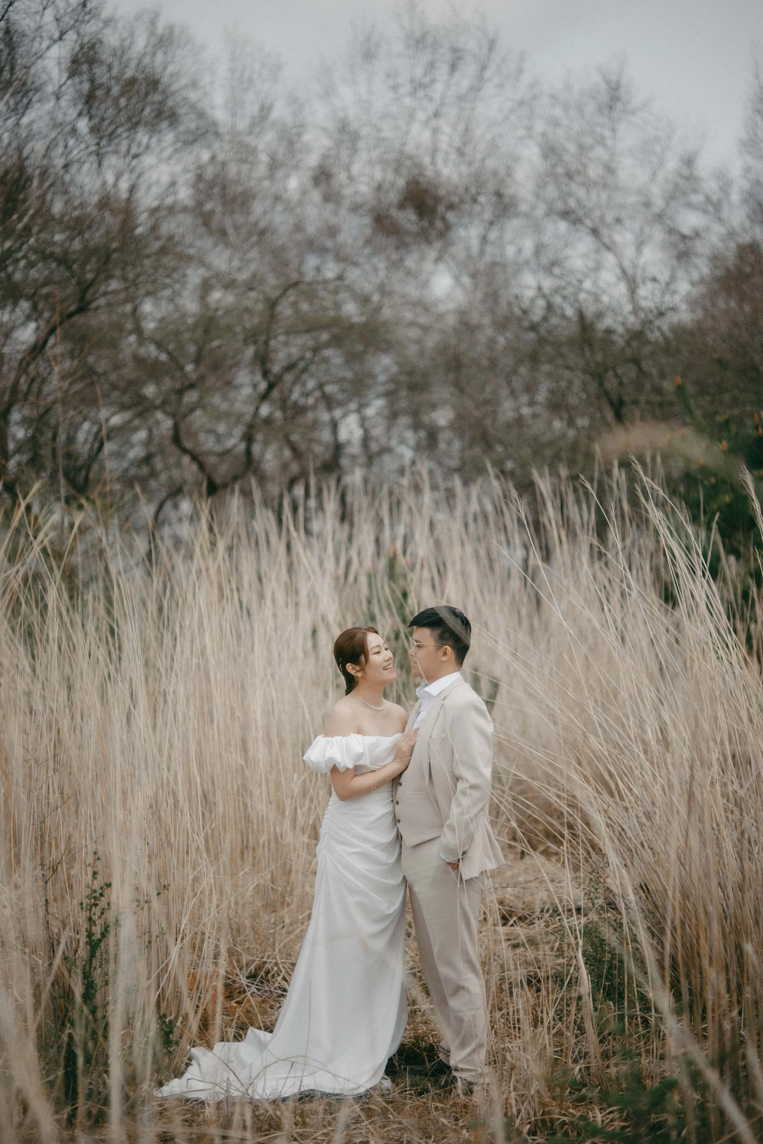 A bride and groom in wedding attire standing face-to-face in a field of tall, beige grasses with leafless trees in the background.