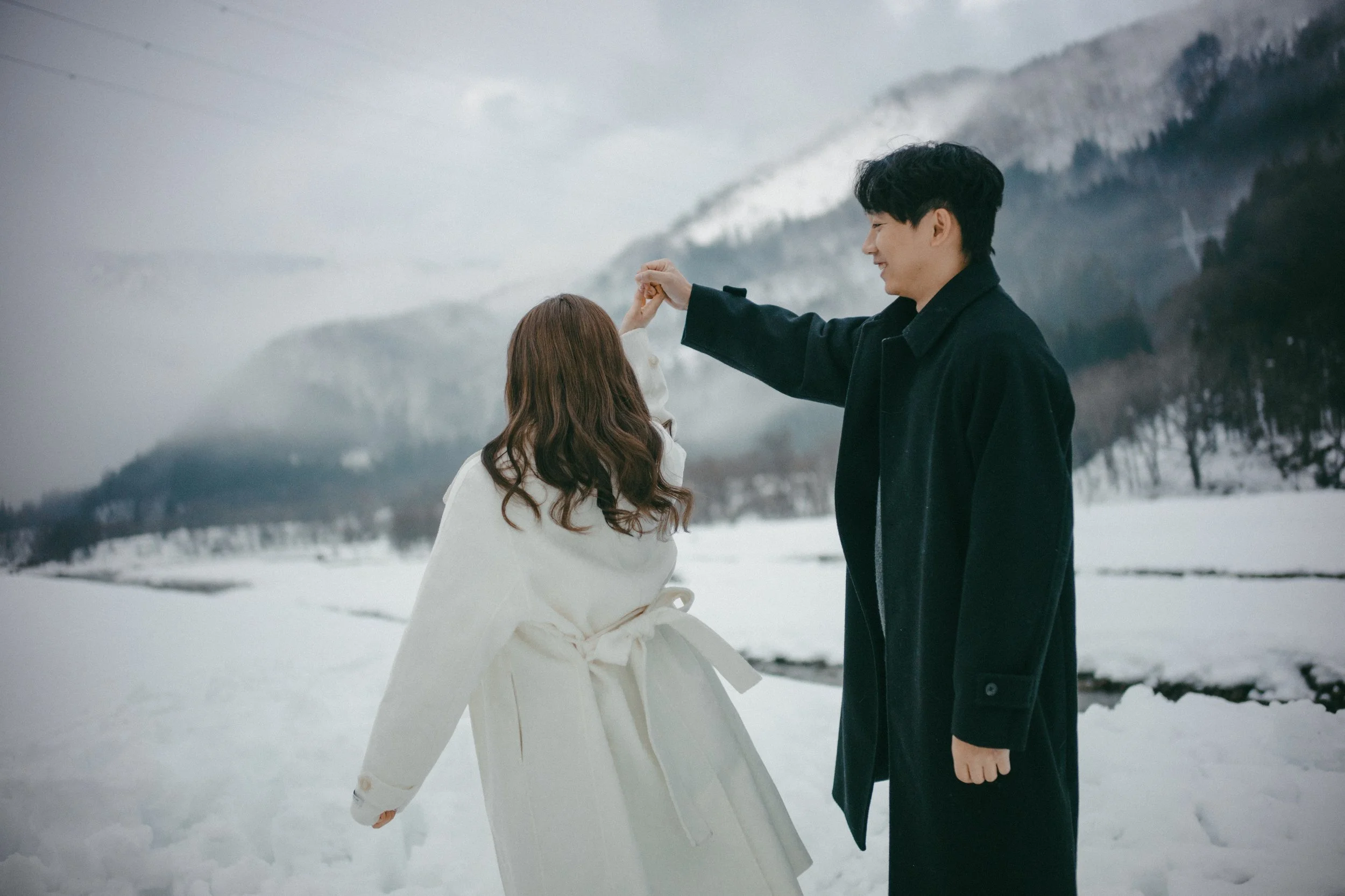 A man twirling a woman in a snowy outdoor landscape with mountains in the background.