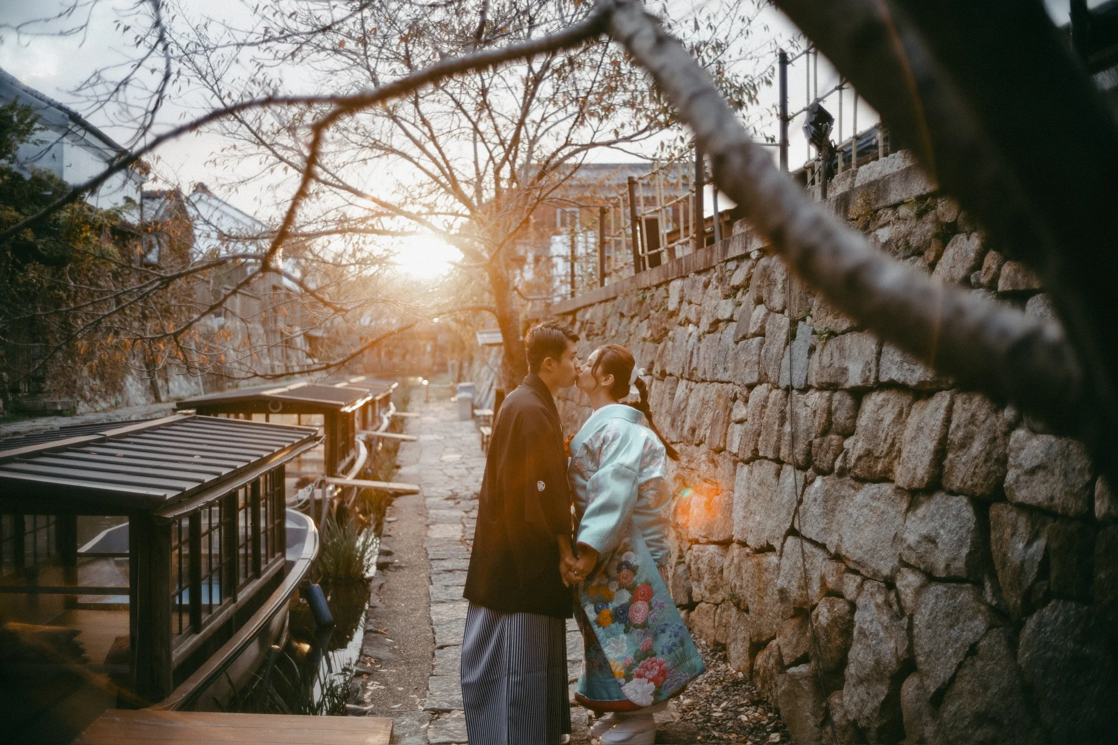 A couple dressed in traditional Japanese attire sharing a kiss on a stone path during sunset, with a stone wall and leafless trees in the background.