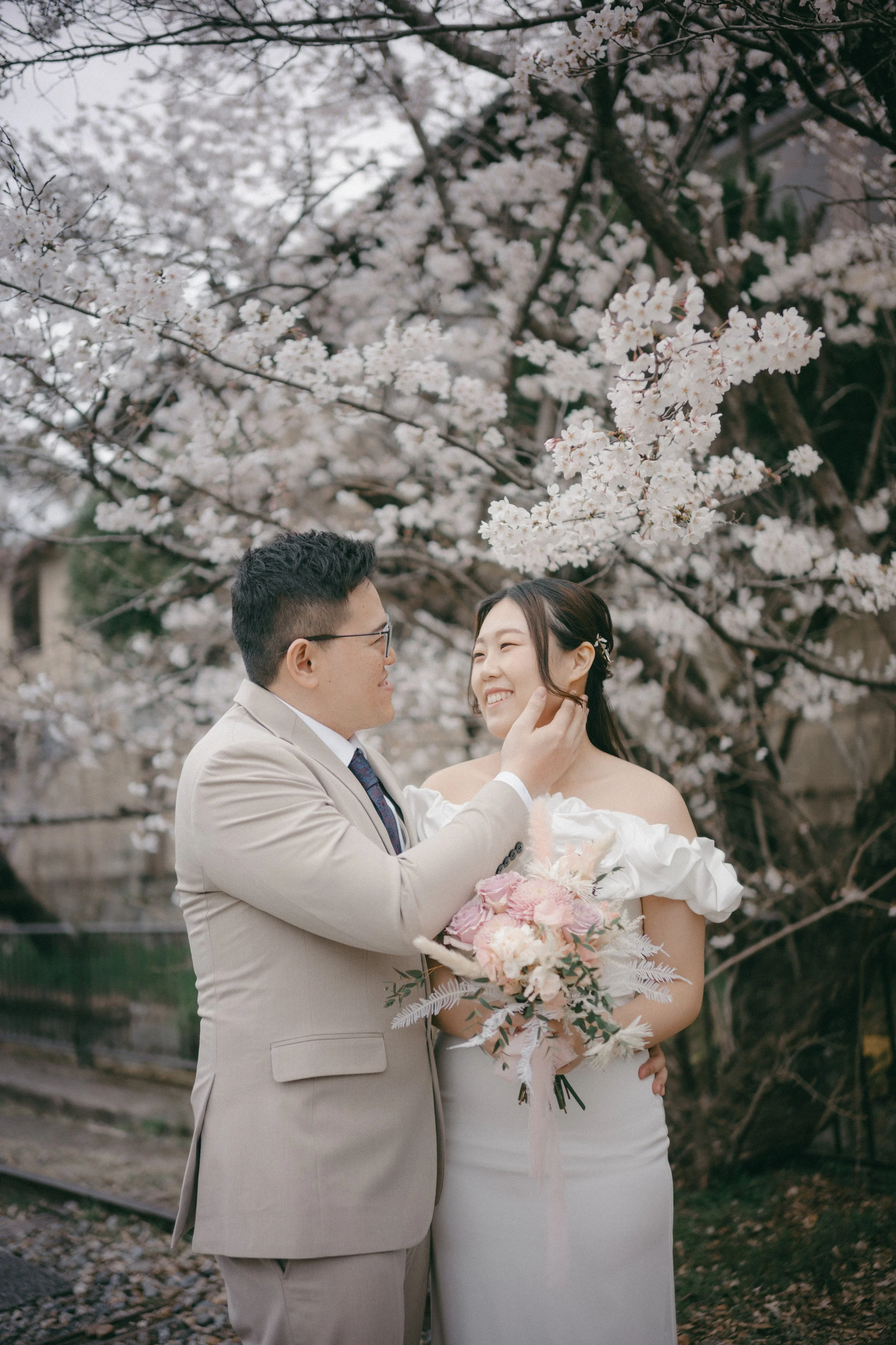 A couple in wedding attire sharing a tender moment under blooming cherry blossom trees. The groom gently touches the bride's face while they smile at each other. The bride holds a bouquet of pink and white flowers.