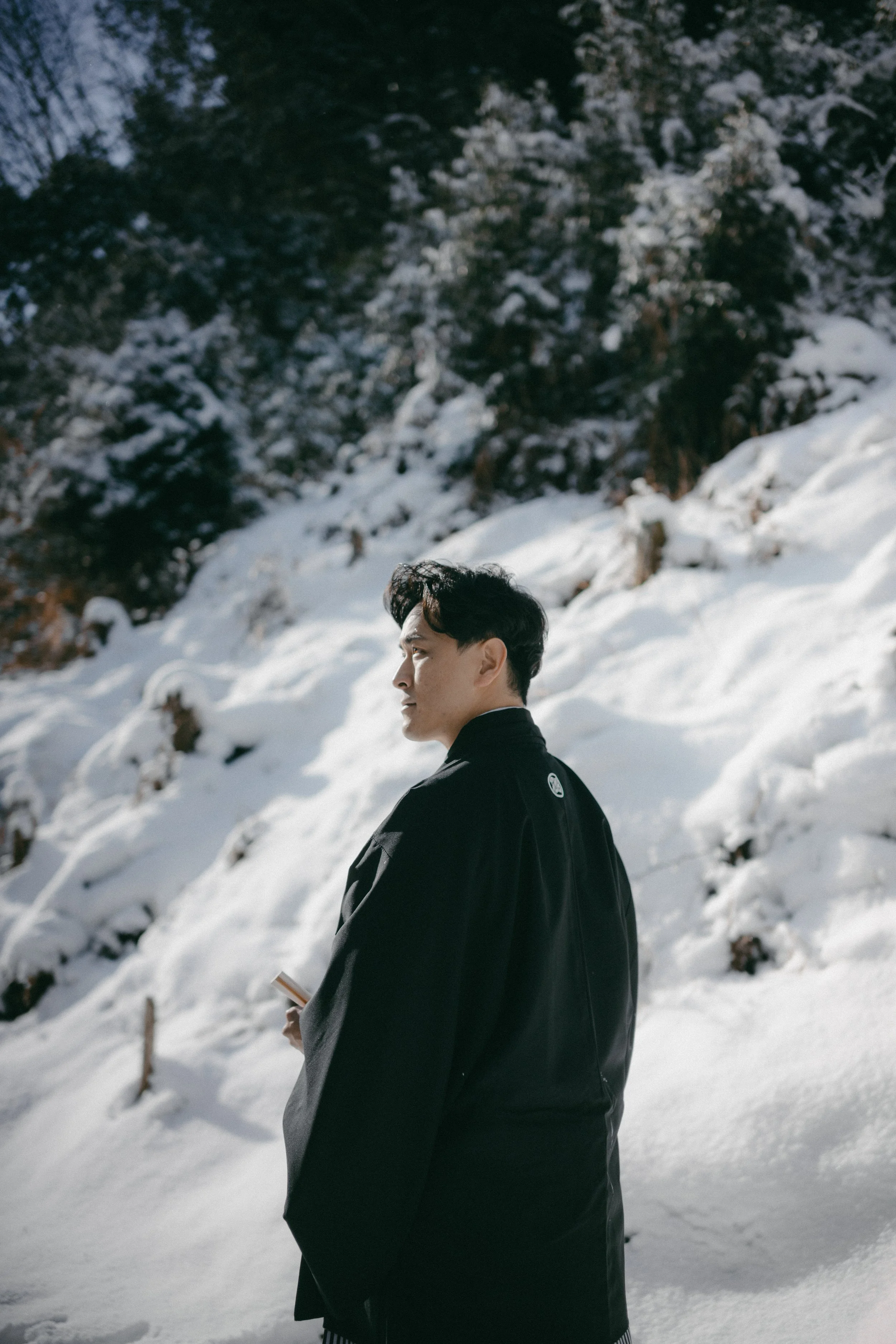 A man dressed in winter clothing stands in a snowy landscape with evergreen trees in the background, looking to the side.