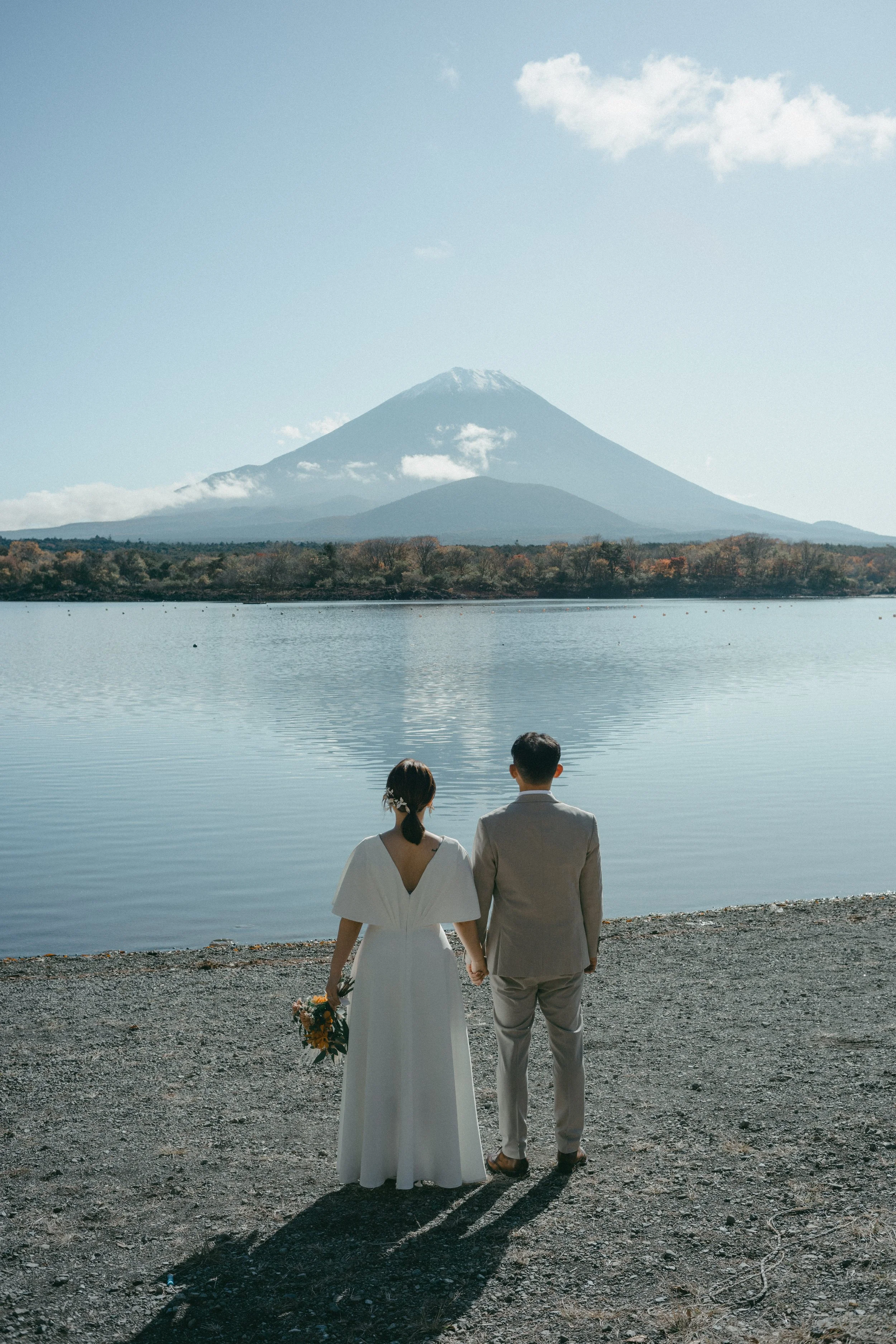 A bride and groom holding hands and facing a lake with Mount Fuji in the background, during daytime.