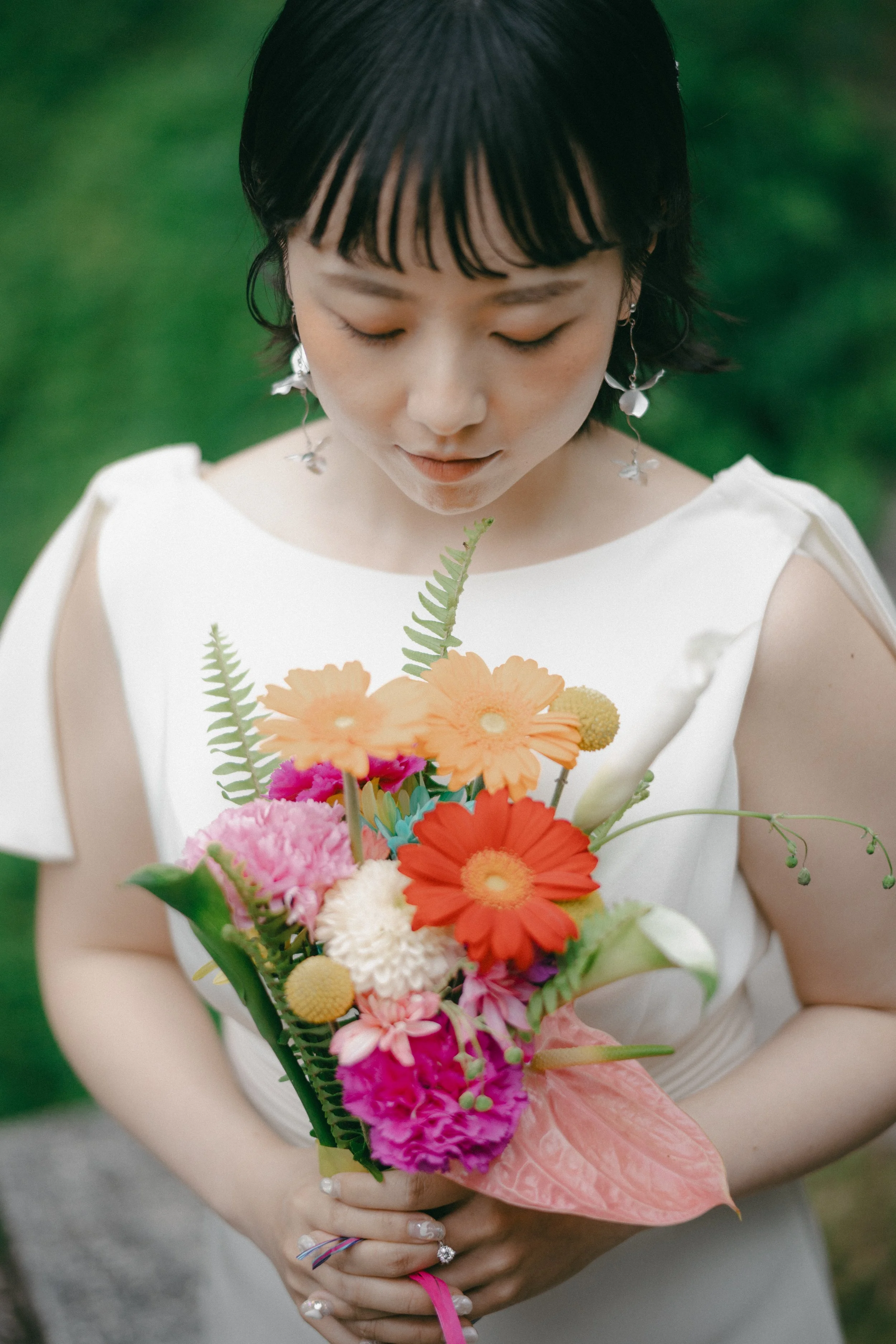Woman in a white dress holding a colorful bouquet of flowers, looking down at it, outdoors with green background.