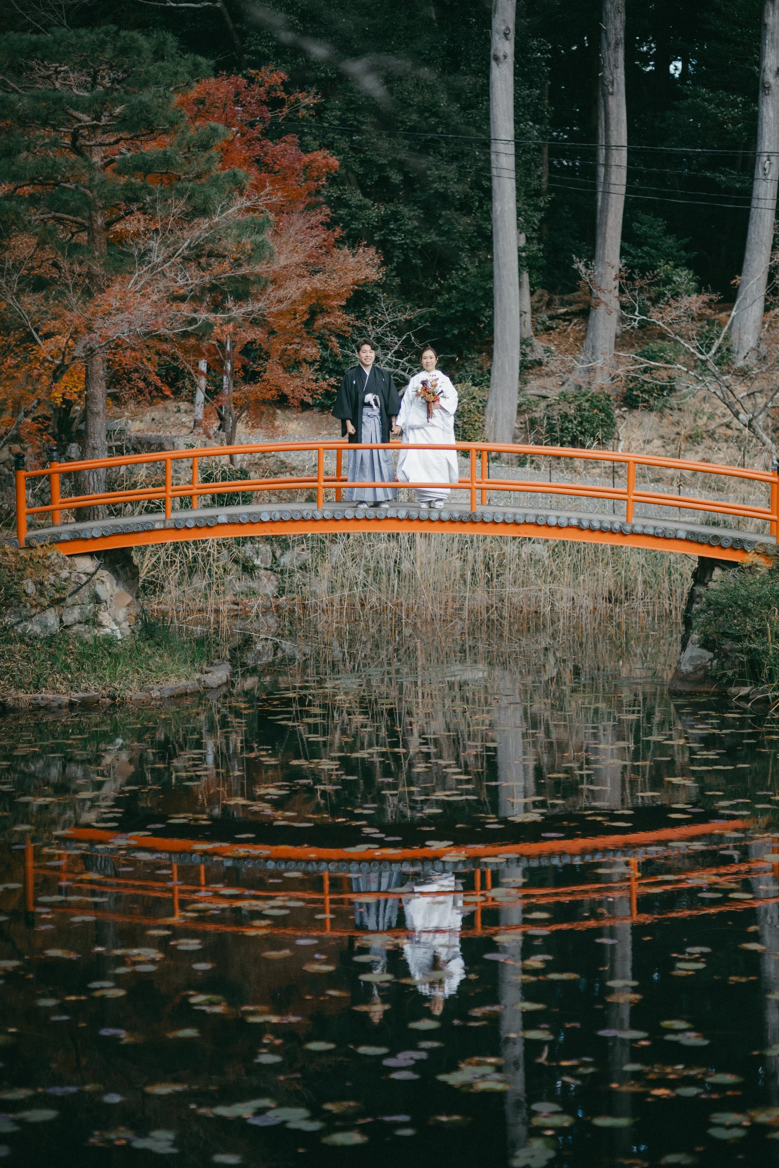 A wedding couple standing on a red bridge over a pond with fall foliage in the background.