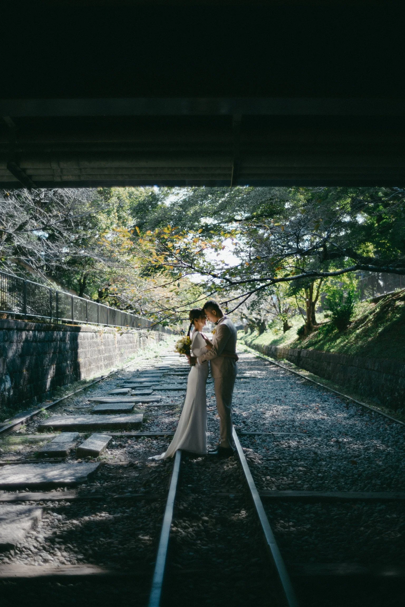 A couple in wedding attire standing close together on railway tracks under a bridge, with trees and sunlight in the background.