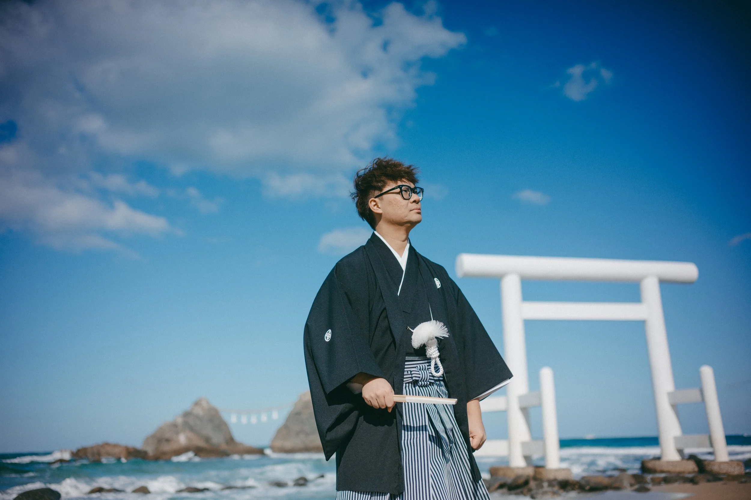 A man in traditional Japanese attire standing on a rocky beach with a torii gate and rocks in the background during daytime.