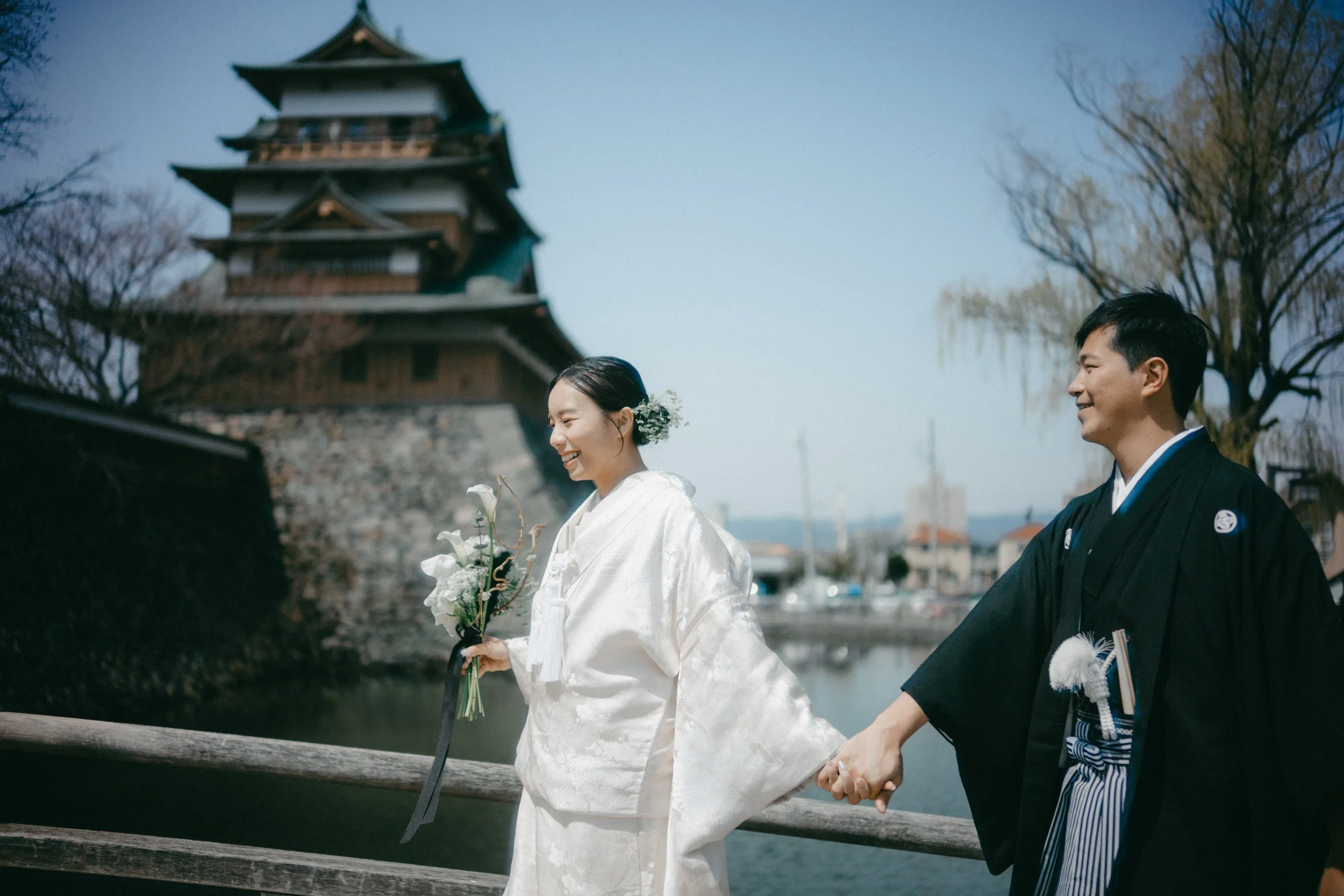A bride and groom in traditional Japanese wedding attire holding hands outside near water with a historic Japanese building and trees in the background, smiling.