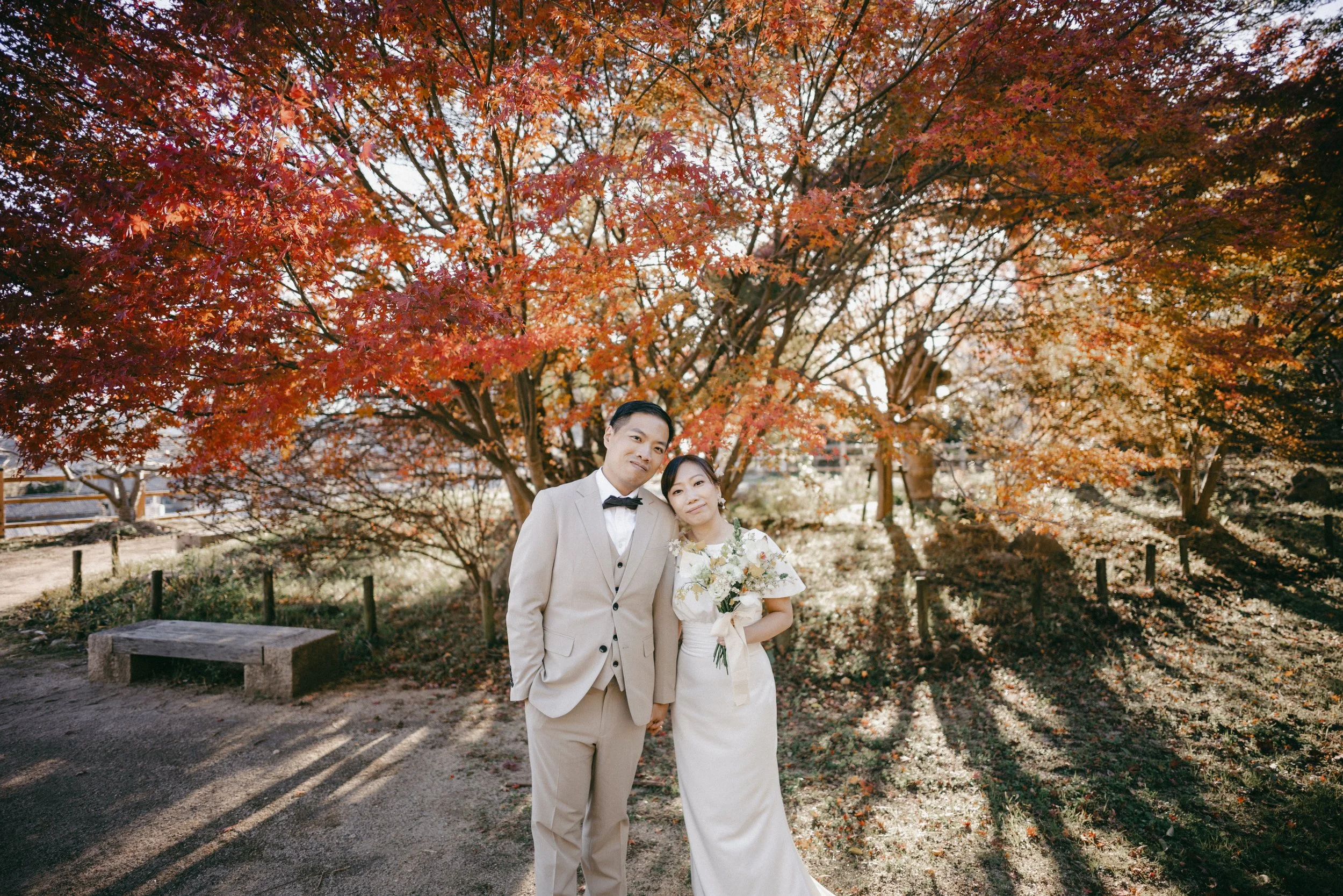 A bride and groom in wedding attire posing outdoors in front of a large, vibrant red and orange autumn tree, with sunlight casting long shadows on the ground.