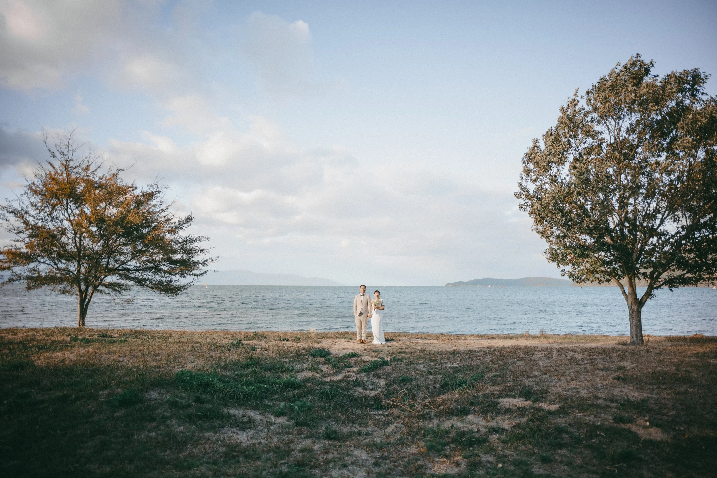 A couple in wedding attire standing by the water on a beach with two trees on either side, cloudy sky, distant mountains.