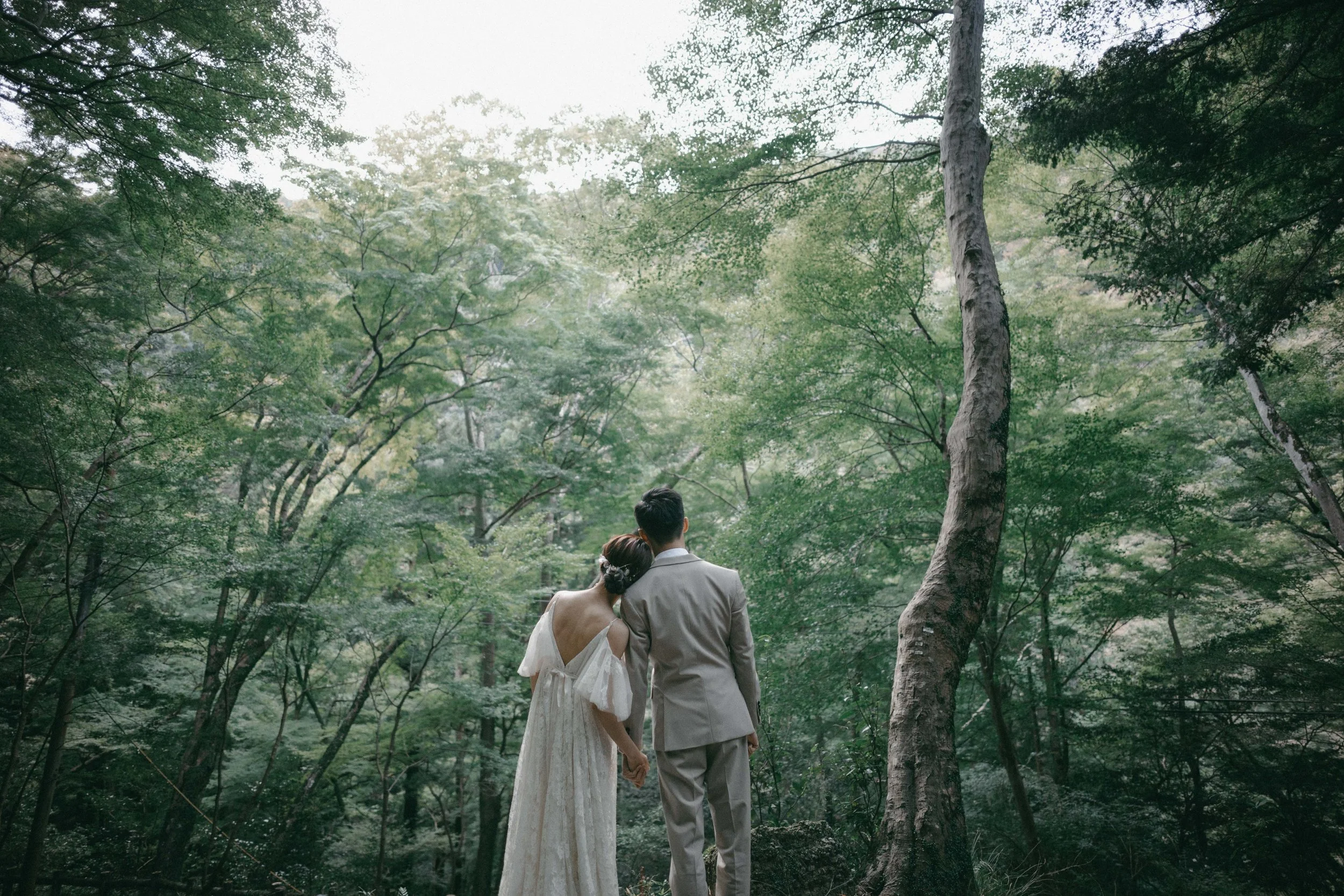 A bride and groom in wedding attire holding hands and walking through a lush green forest.
