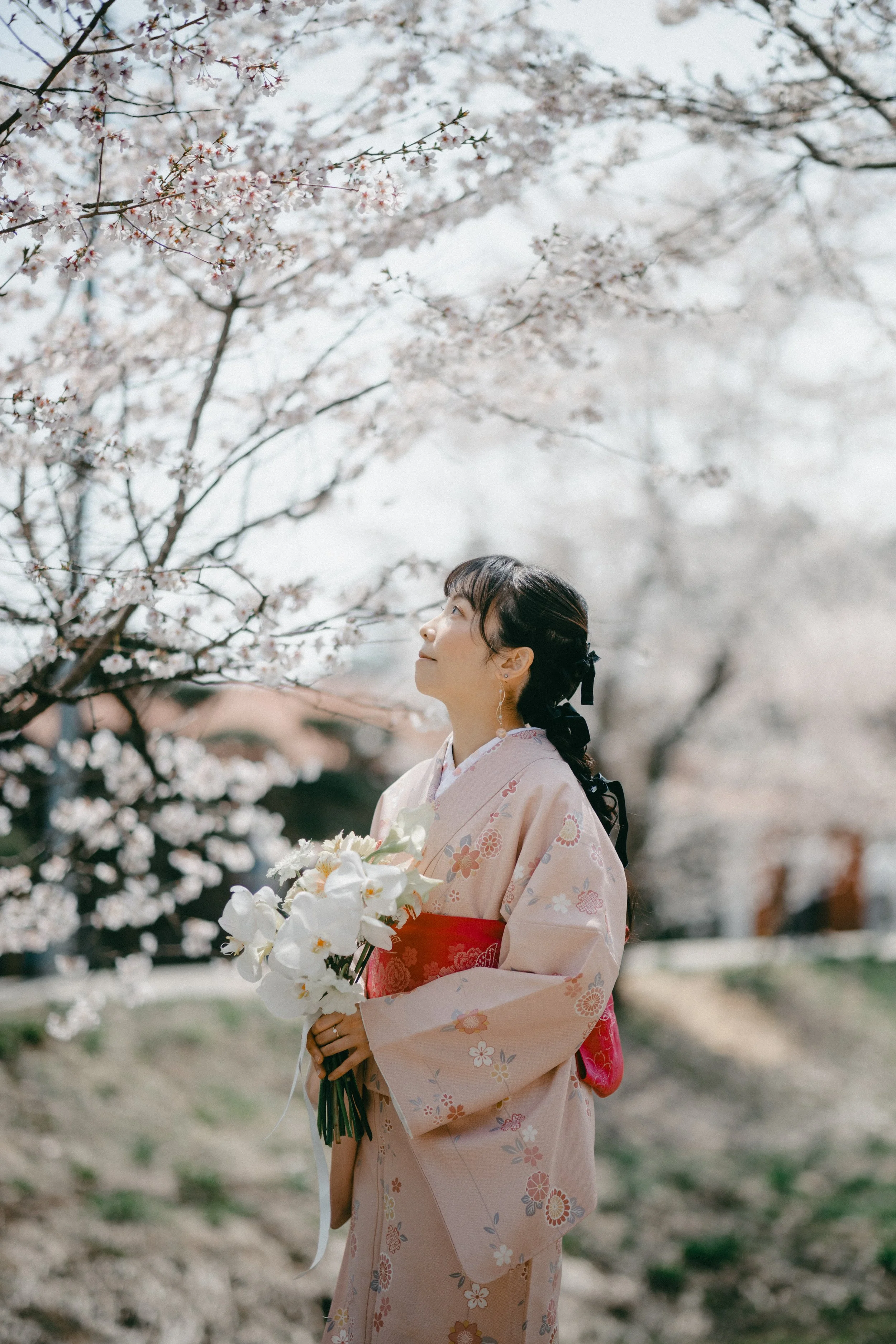 A woman in a traditional beige kimono with pink and floral designs stands under blooming cherry blossom trees, holding a bouquet of white flowers, and gazing upwards at the blossoms.
