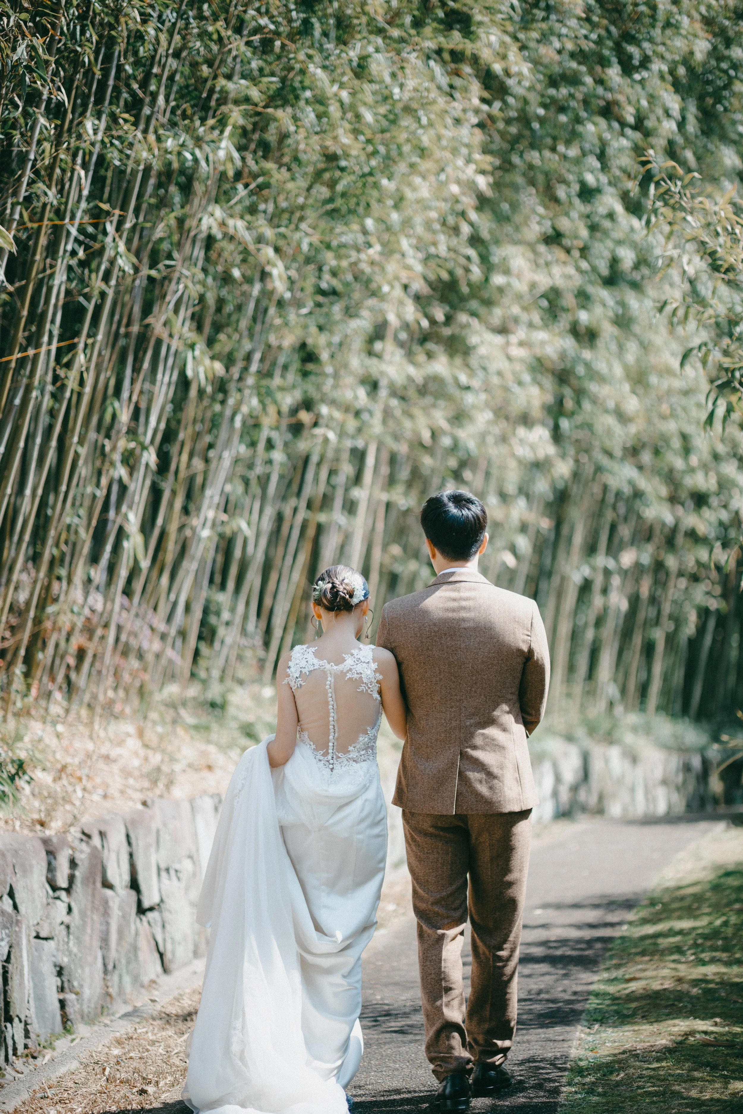 A bride and groom walking together on a path surrounded by tall bamboo plants, with the bride wearing a white wedding dress and the groom in a brown suit.