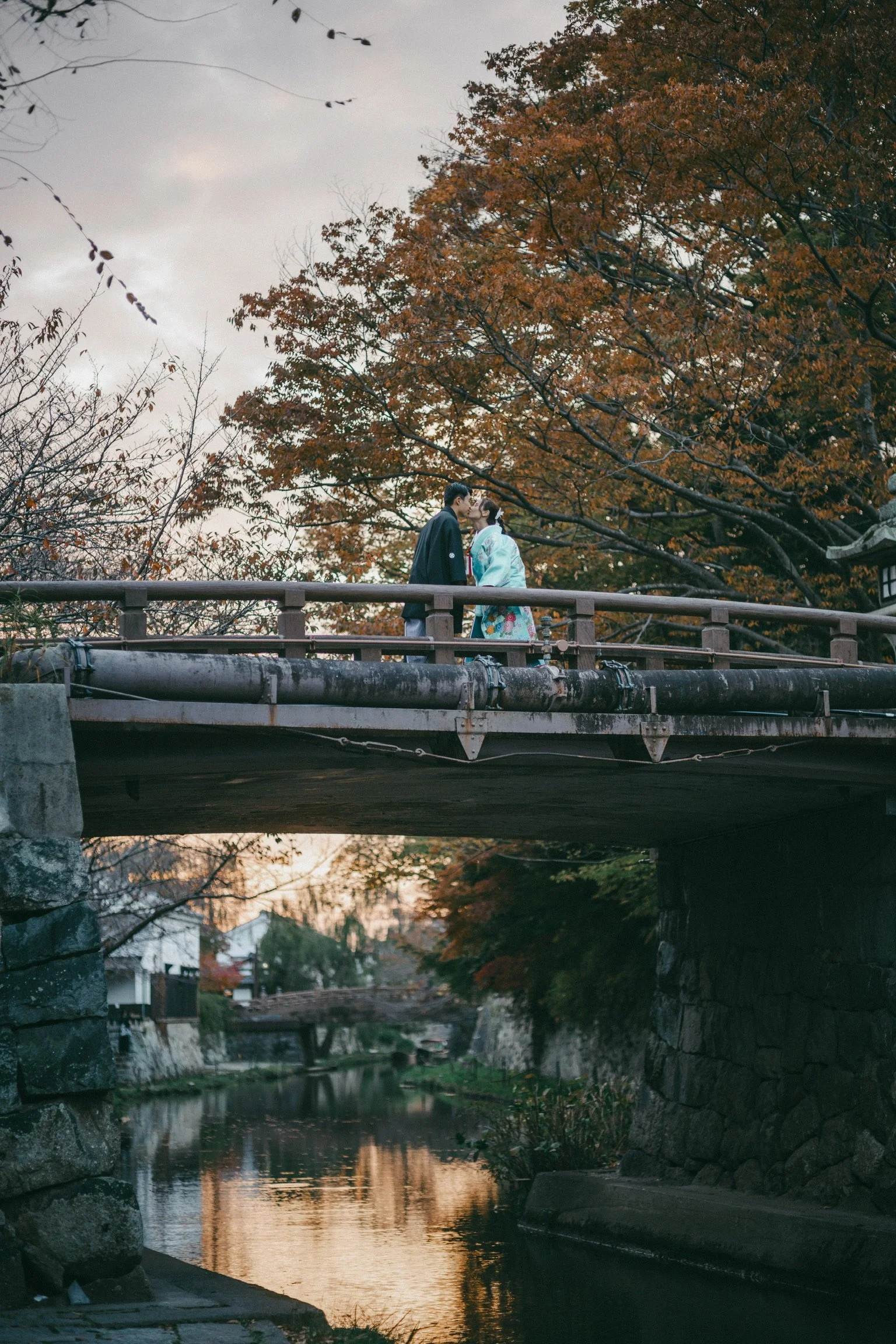 A couple dressed in traditional Japanese kimono sharing a kiss on a bridge over a river during autumn, with orange and yellow trees surrounding them.