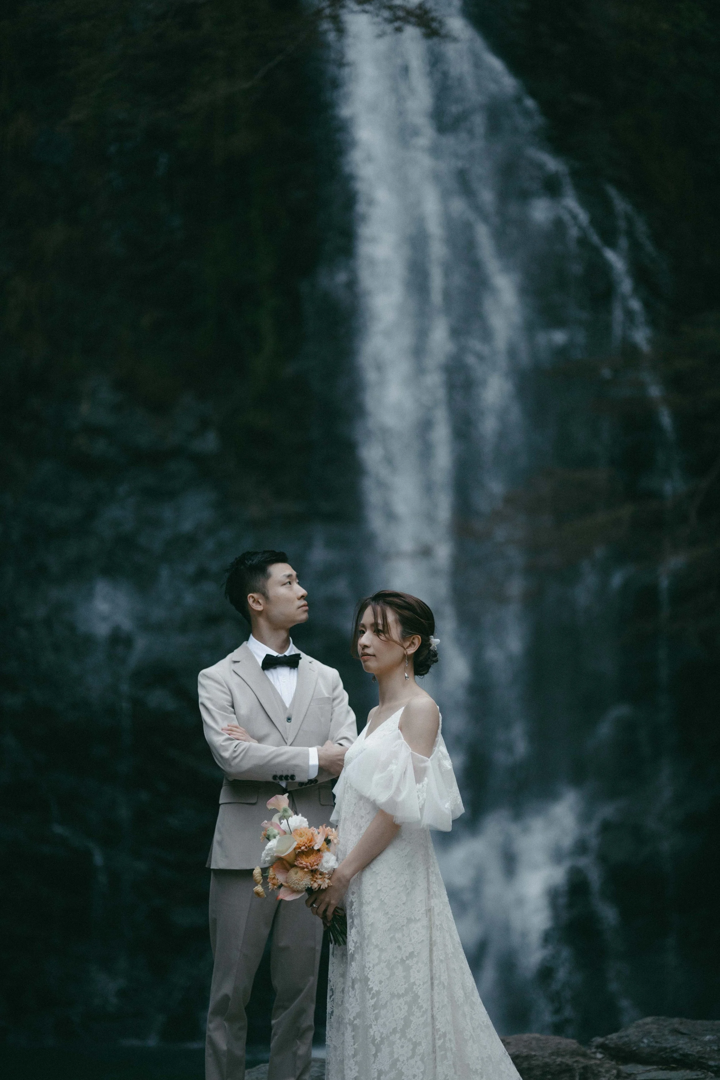 A bride and groom standing in front of a waterfall, the groom in a light suit with a bow tie and the bride in a lace wedding dress holding a bouquet of flowers.