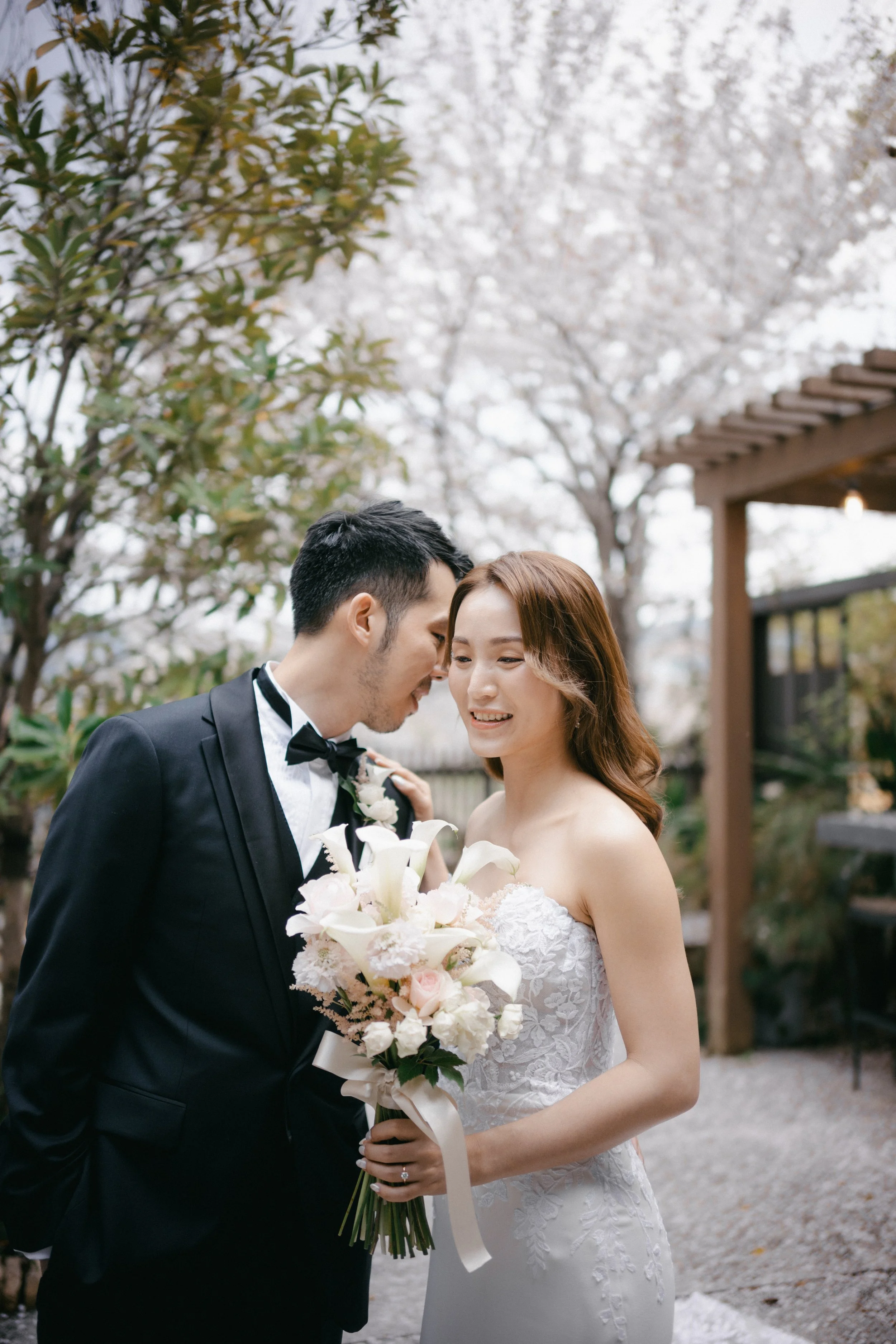 A bride and groom on their wedding day, standing outdoors among trees blooming with white flowers. The bride is smiling and holding a bouquet of white and pink flowers, wearing a white strapless wedding gown. The groom is leaning towards her, wearing