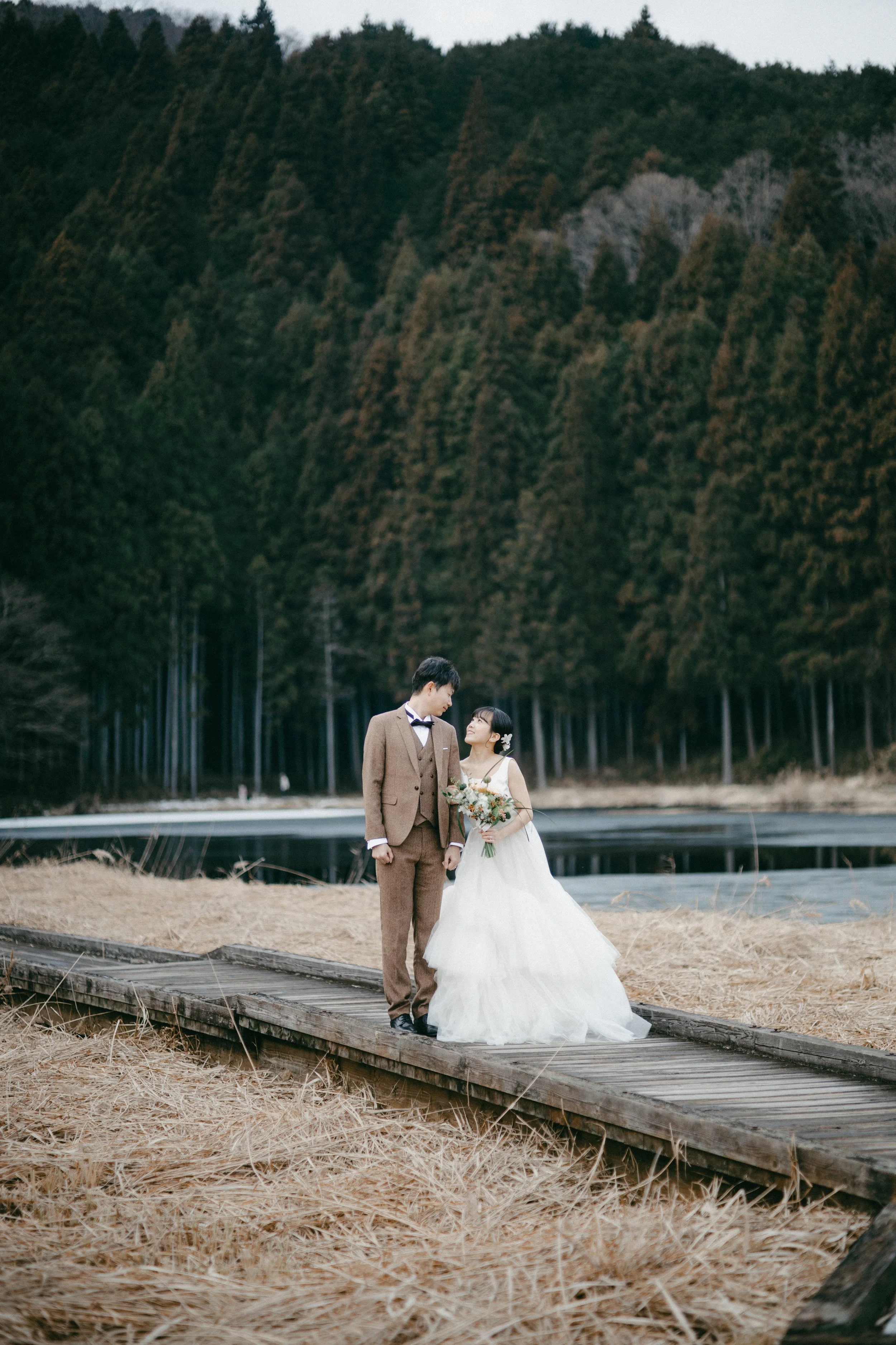 A bride and groom standing on a wooden pathway by a lake, surrounded by trees, with mountains in the background, dressed in wedding attire.