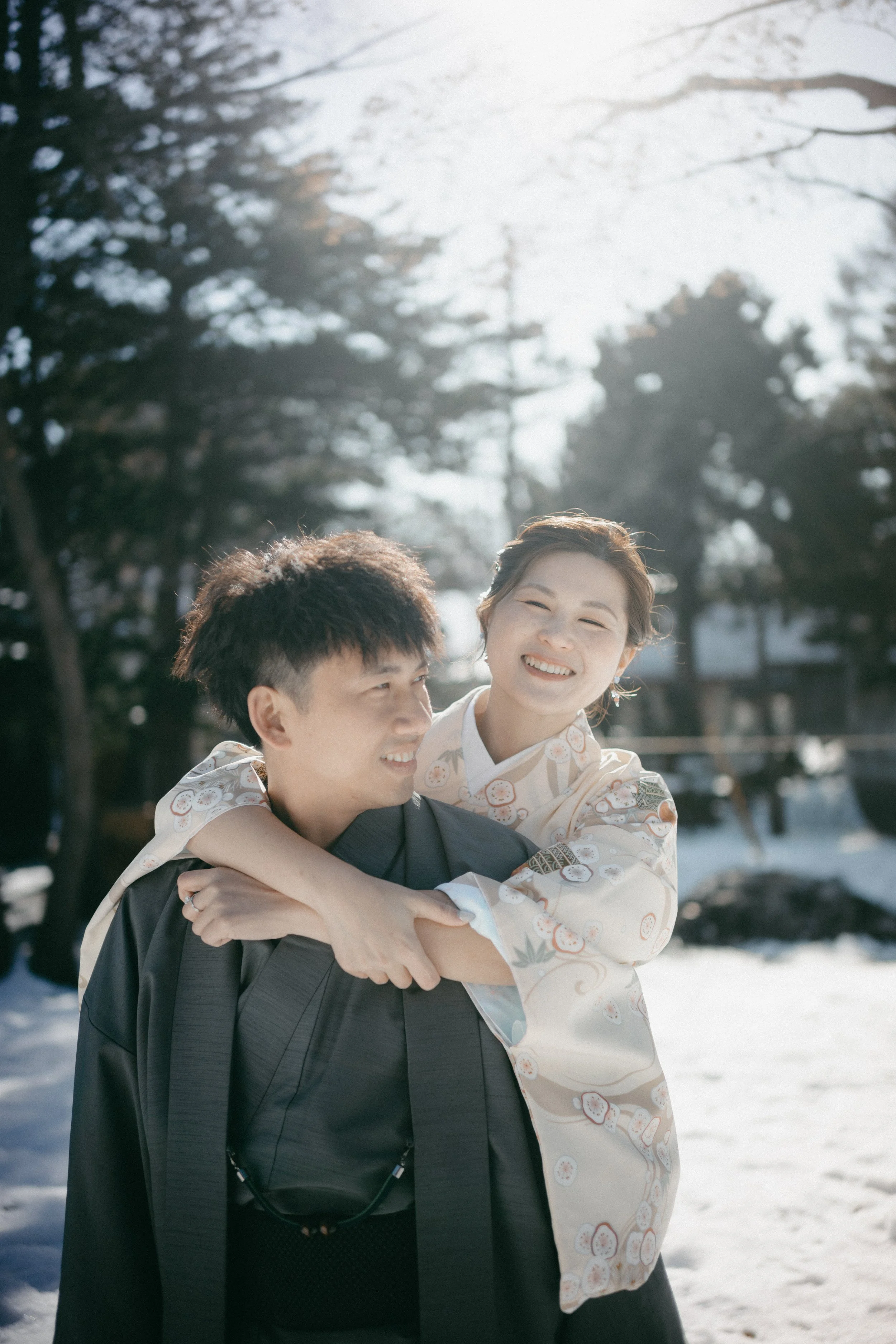 A smiling woman in a traditional Japanese kimono hugging a man in a black kimono outdoors in a snowy landscape with trees.