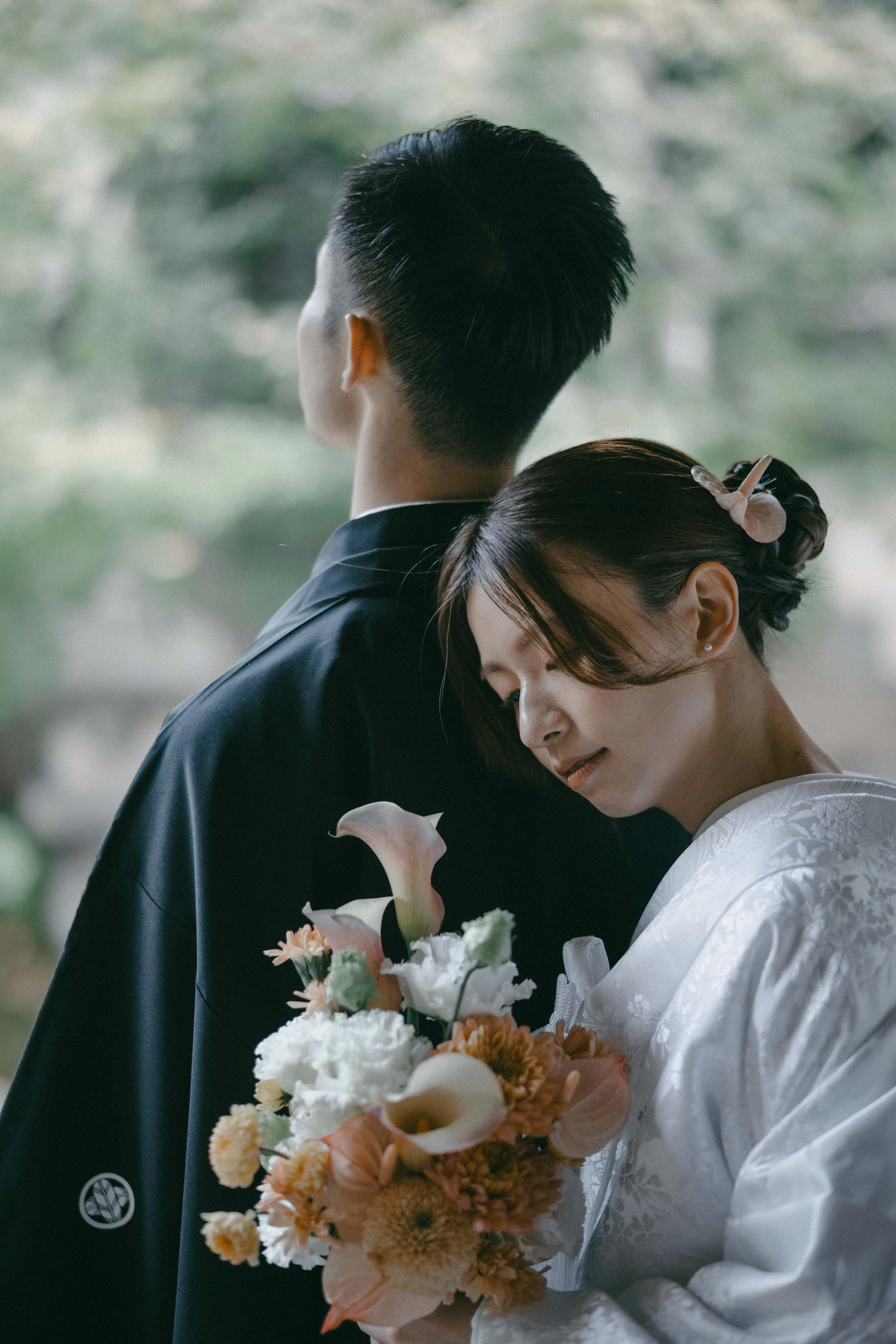 A woman in a traditional white wedding kimono holding a bouquet of flowers, leaning her head on a man in a black suit, with her eyes closed and a gentle smile.