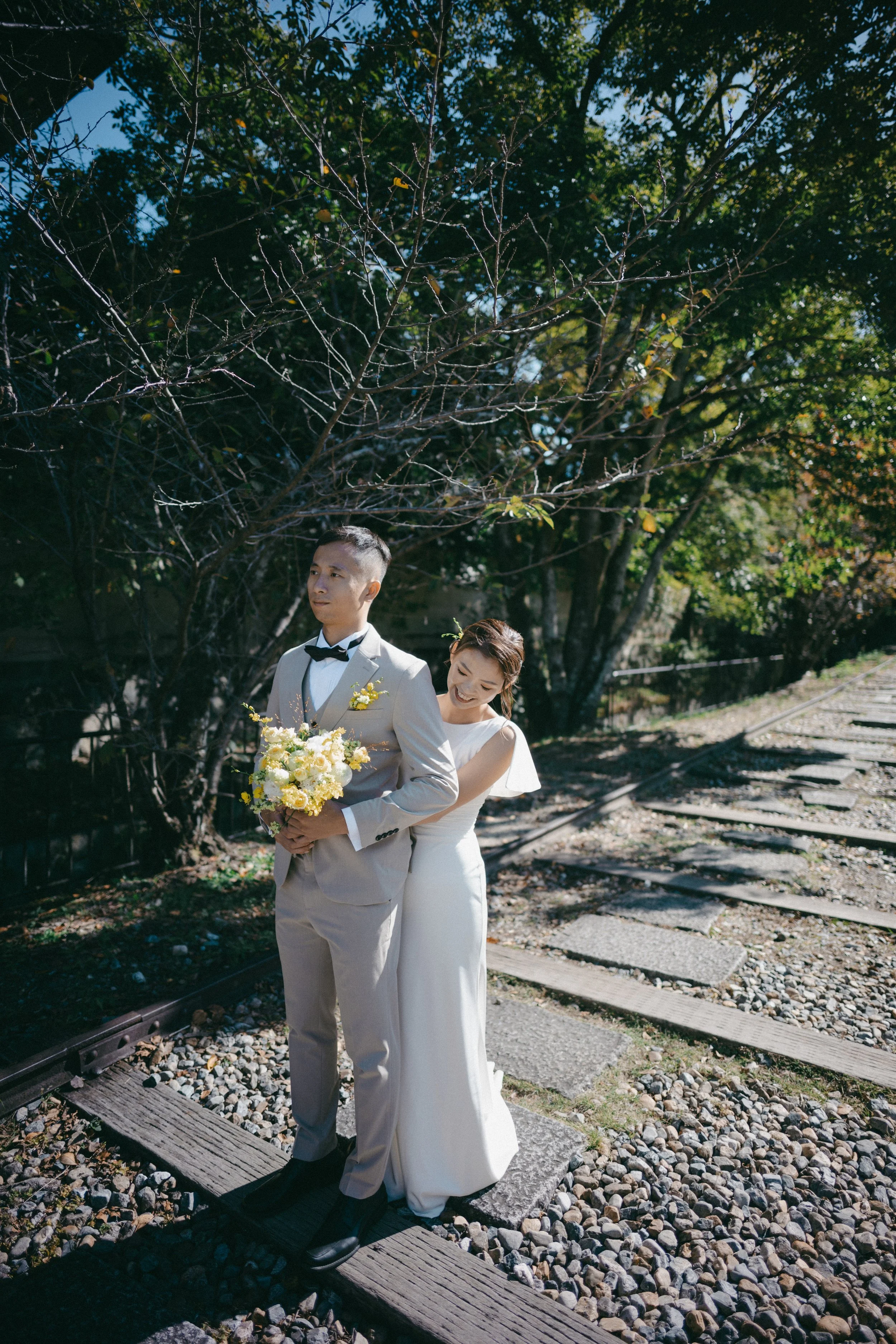 A bride and groom standing on train tracks, with the groom holding a bouquet of yellow and white flowers. The bride is standing behind the groom, smiling and leaning her head on his shoulder. They are outdoors under a tree with green leaves, and the 