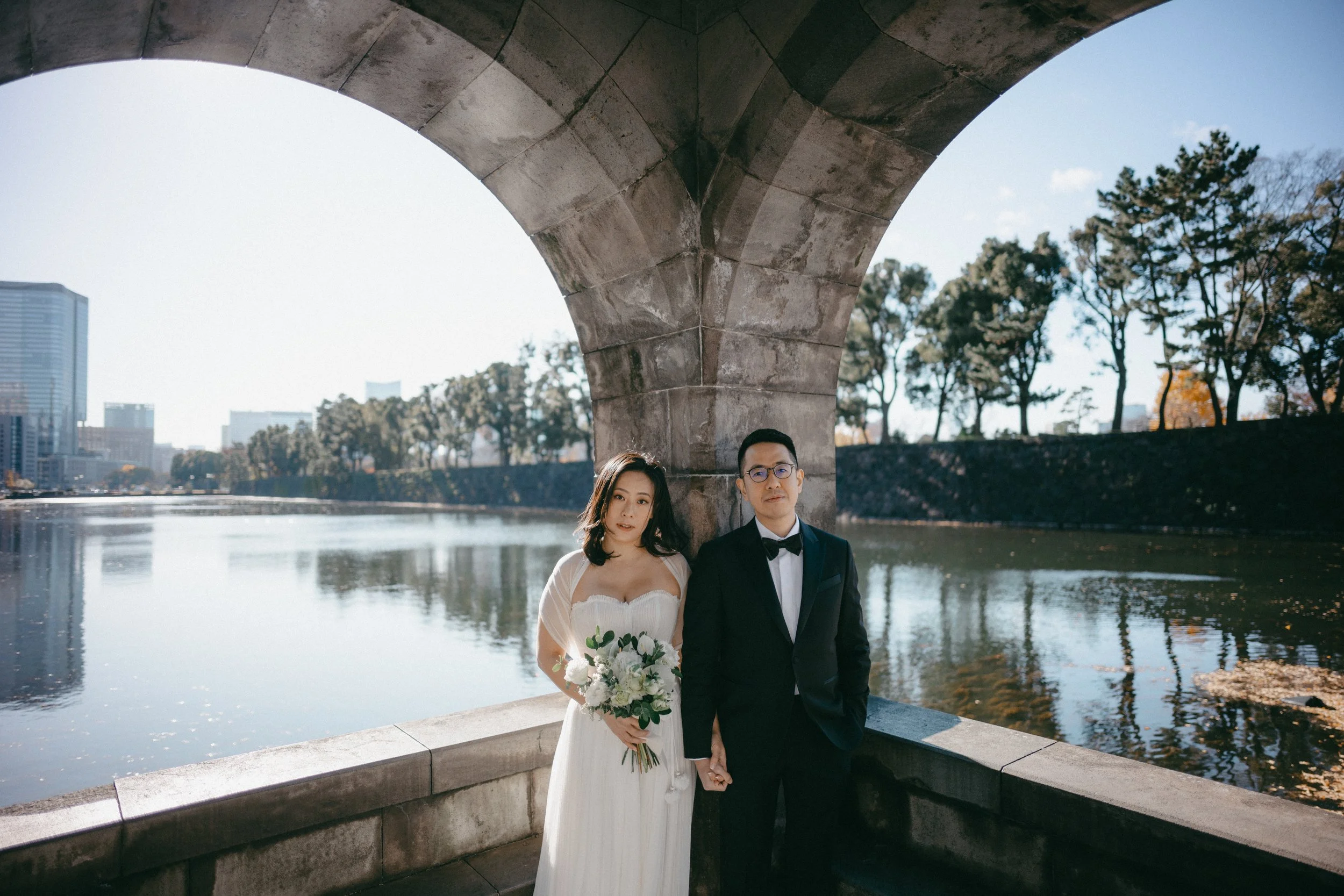 A bride and groom standing hand in hand beneath a stone bridge near a river, with trees and city buildings in the background on a sunny day.