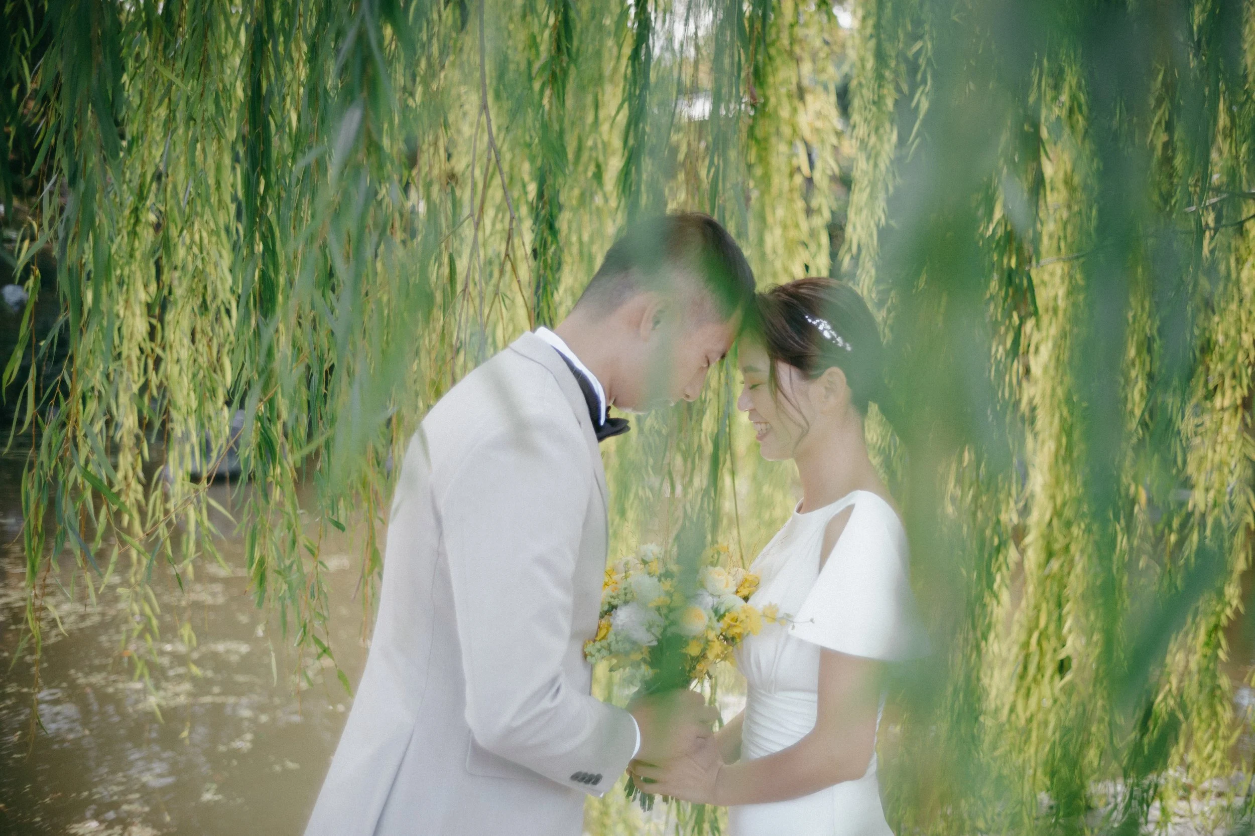 A bride and groom standing under a tree with hanging branches, touching foreheads and smiling, with the bride holding a yellow and white bouquet.