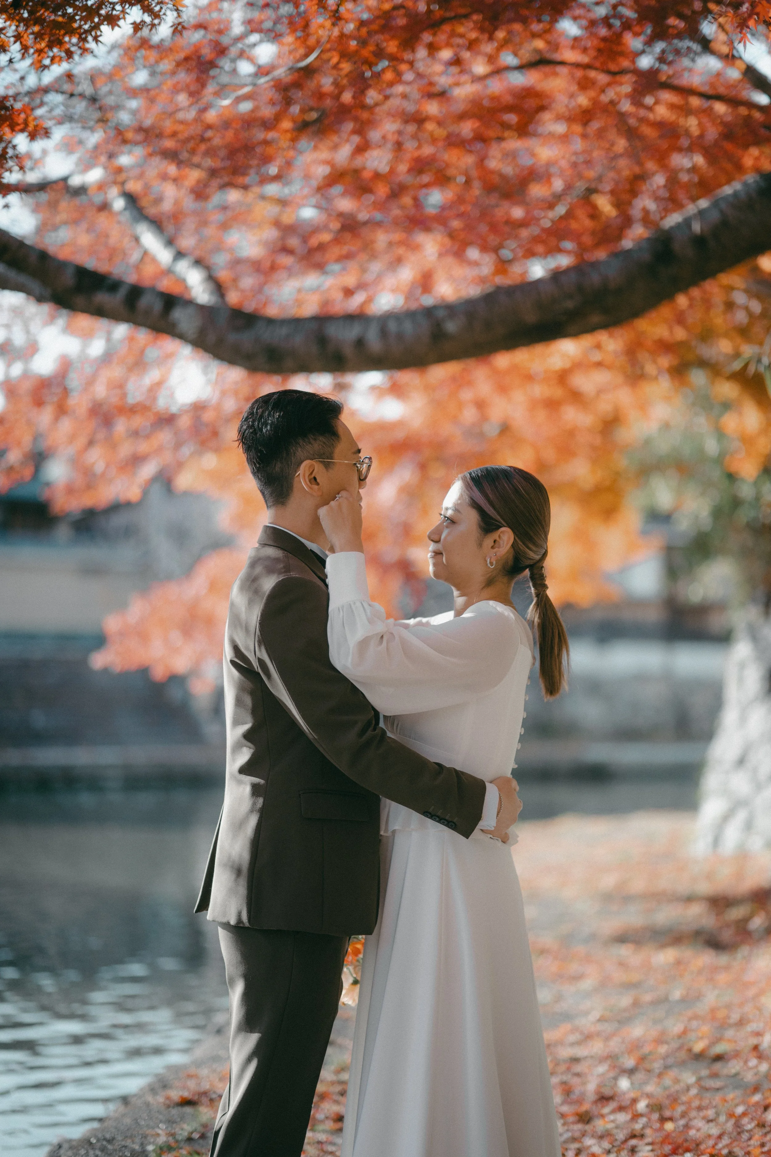 A couple dressed in wedding attire standing close by a body of water surrounded by trees with orange and red autumn leaves, sharing a tender moment.
