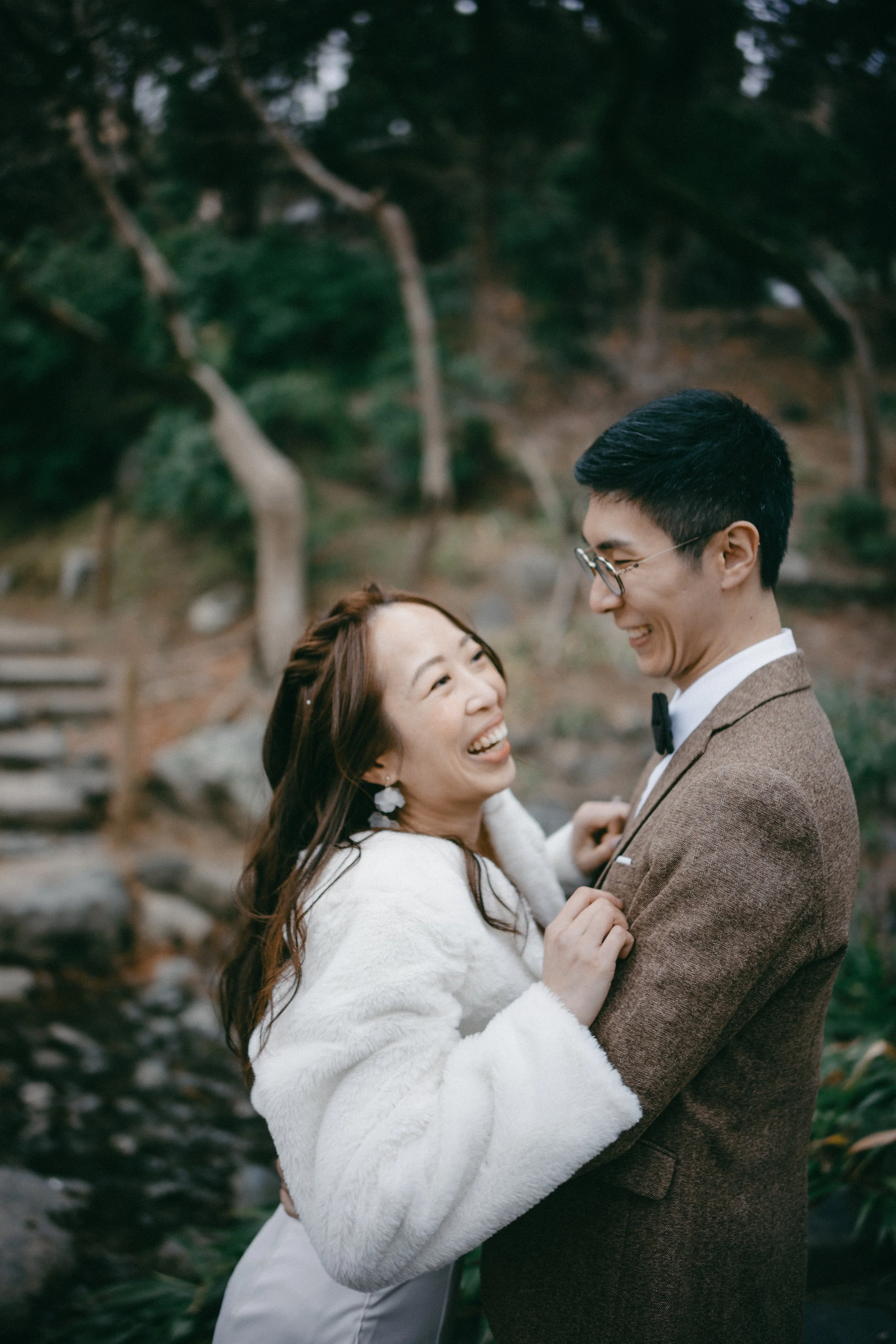 A couple, a woman with long brown hair and a man with glasses, sharing a joyful moment outdoors, smiling and embracing each other in a natural setting with trees and rocks.