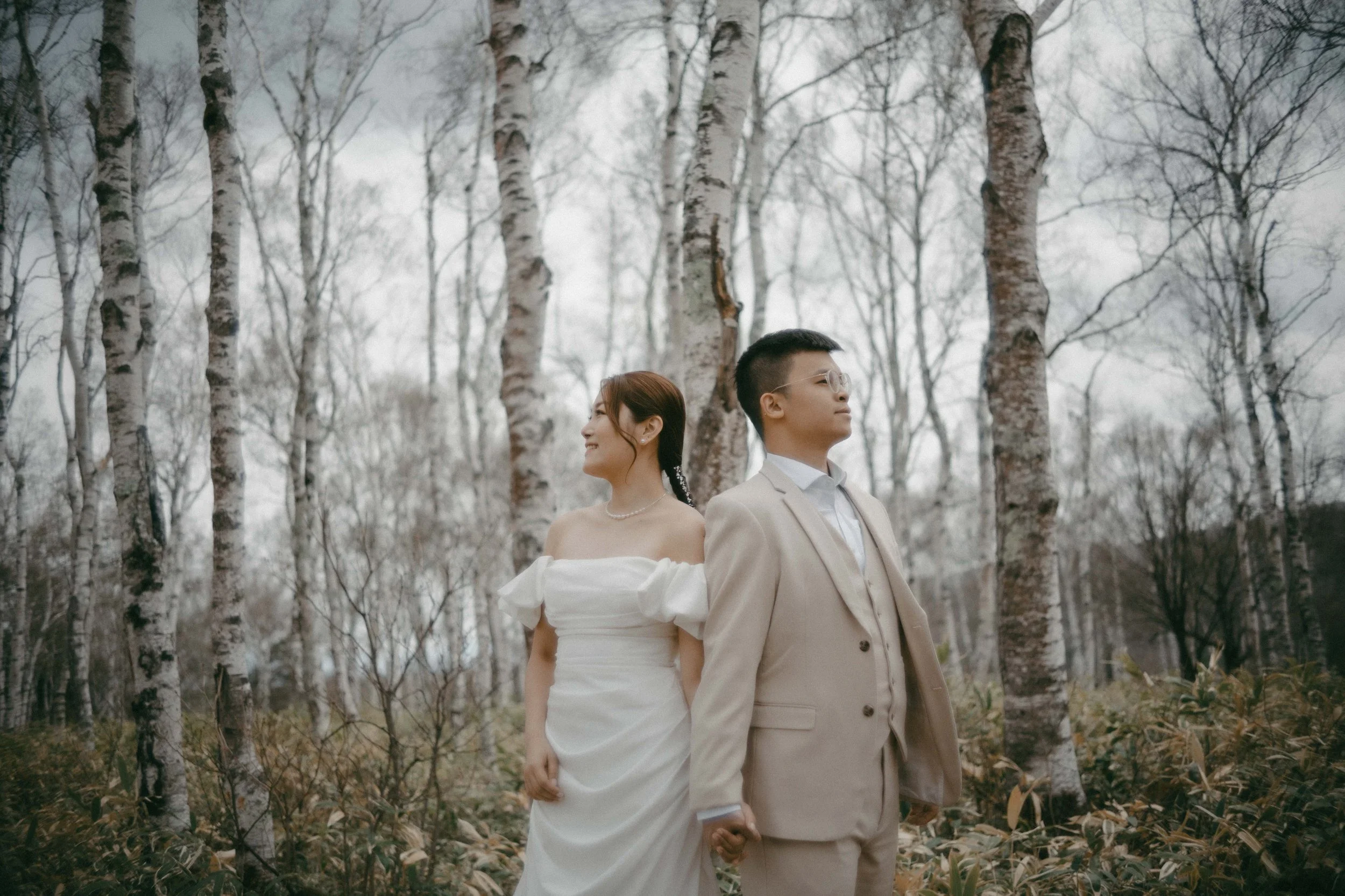 A bride and groom standing back to back in a forest, holding hands, smiling, with bare trees and an overcast sky in the background.