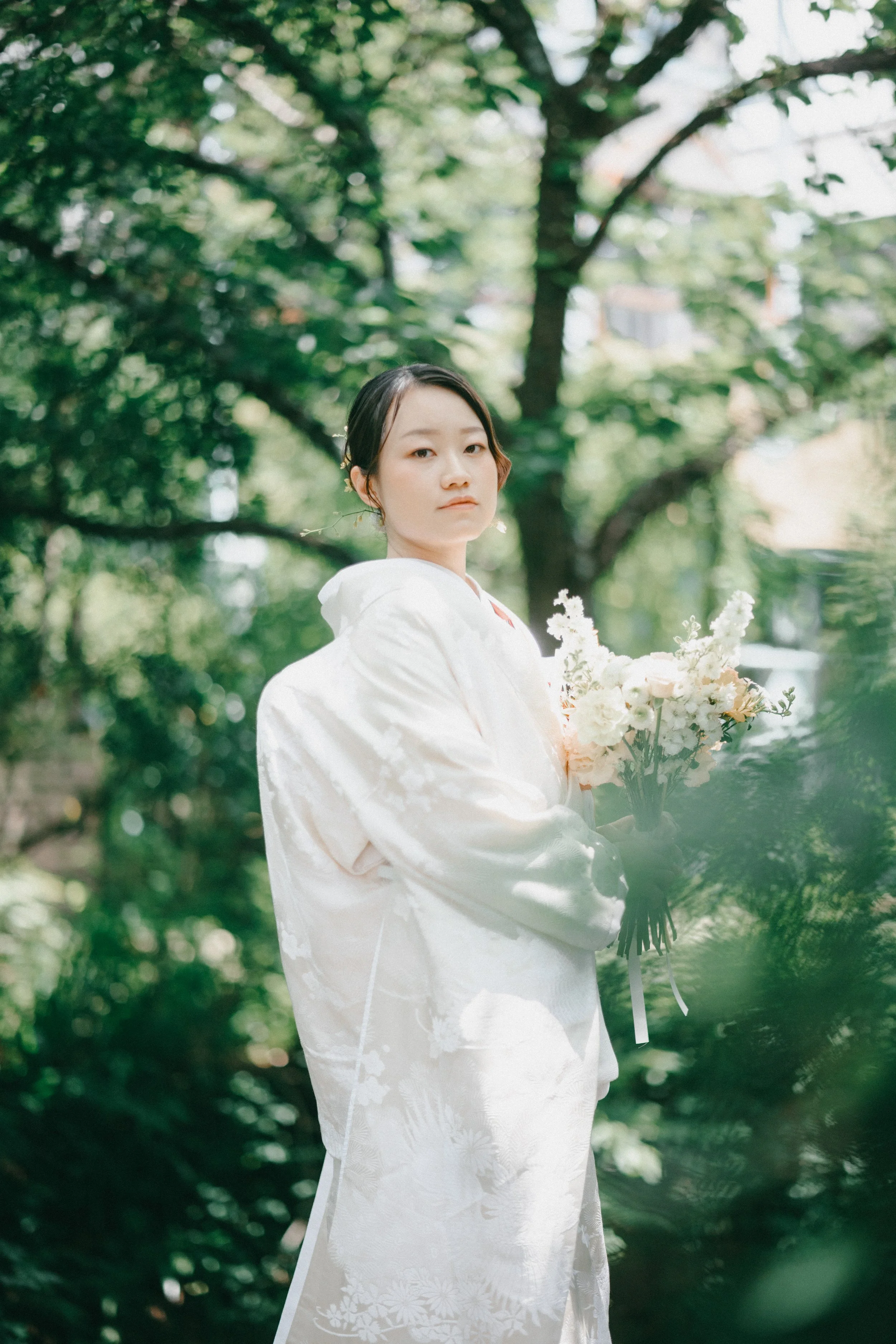A woman in traditional white wedding attire holding a bouquet of white flowers in an outdoor, green, wooded setting.