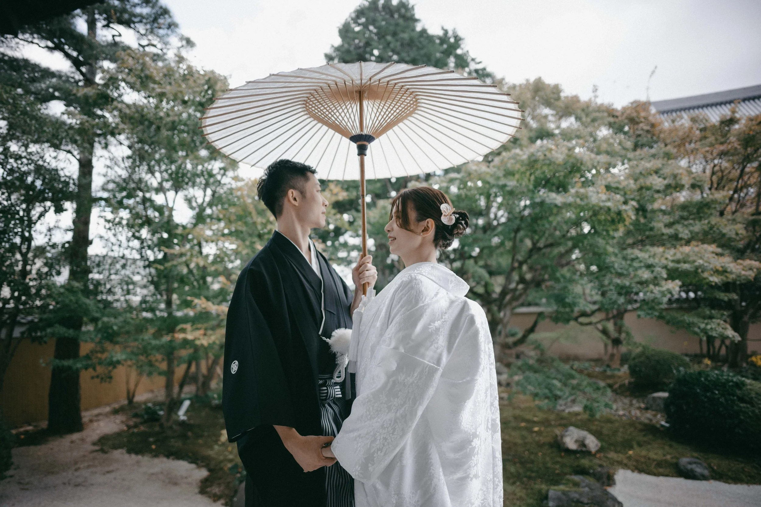 A couple dressed in traditional Japanese wedding attire standing outdoors under a large paper umbrella, gazing into each other's eyes, surrounded by a garden with trees and rocks.