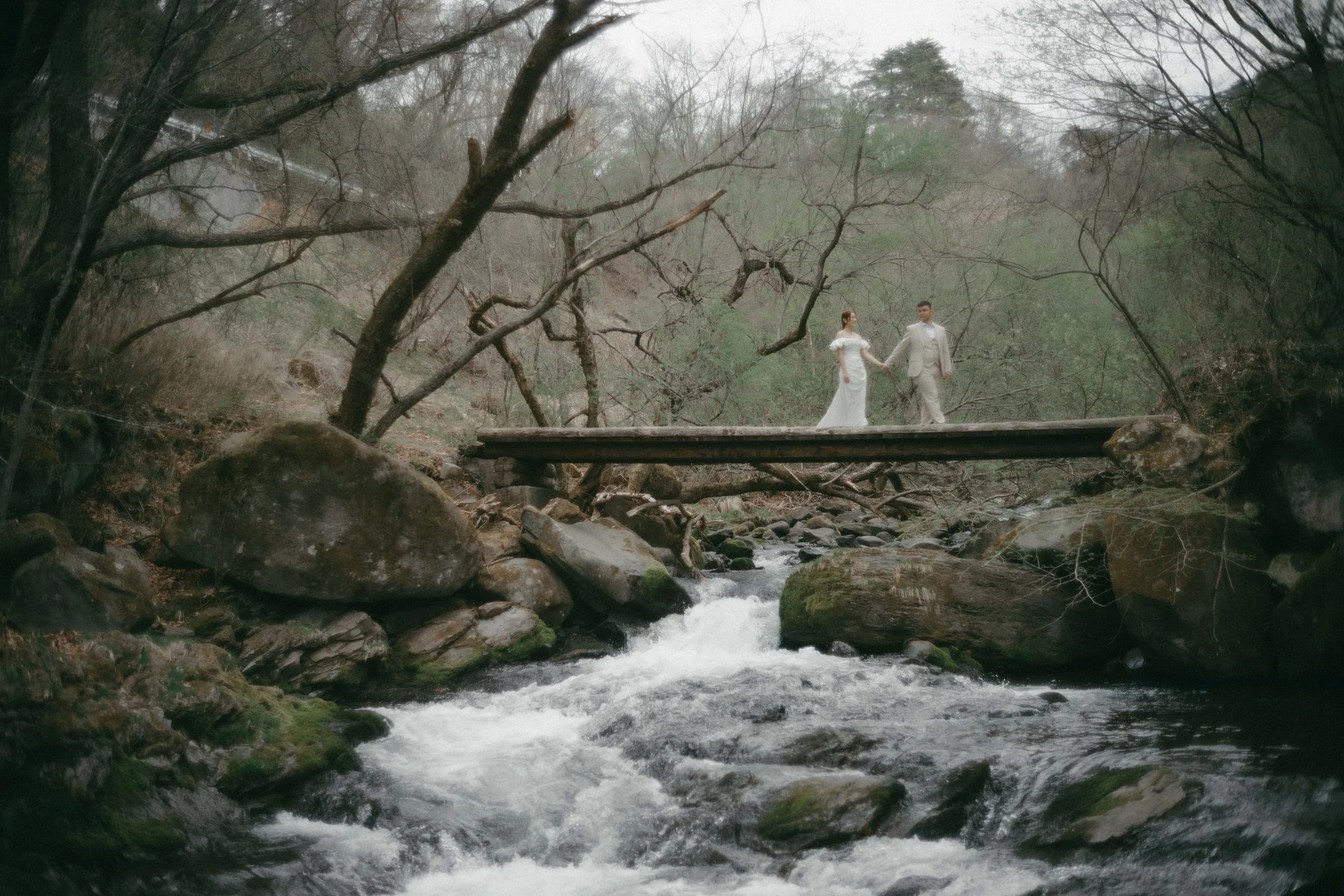A couple in wedding attire standing on a narrow wooden bridge over a rocky stream in a wooded area.