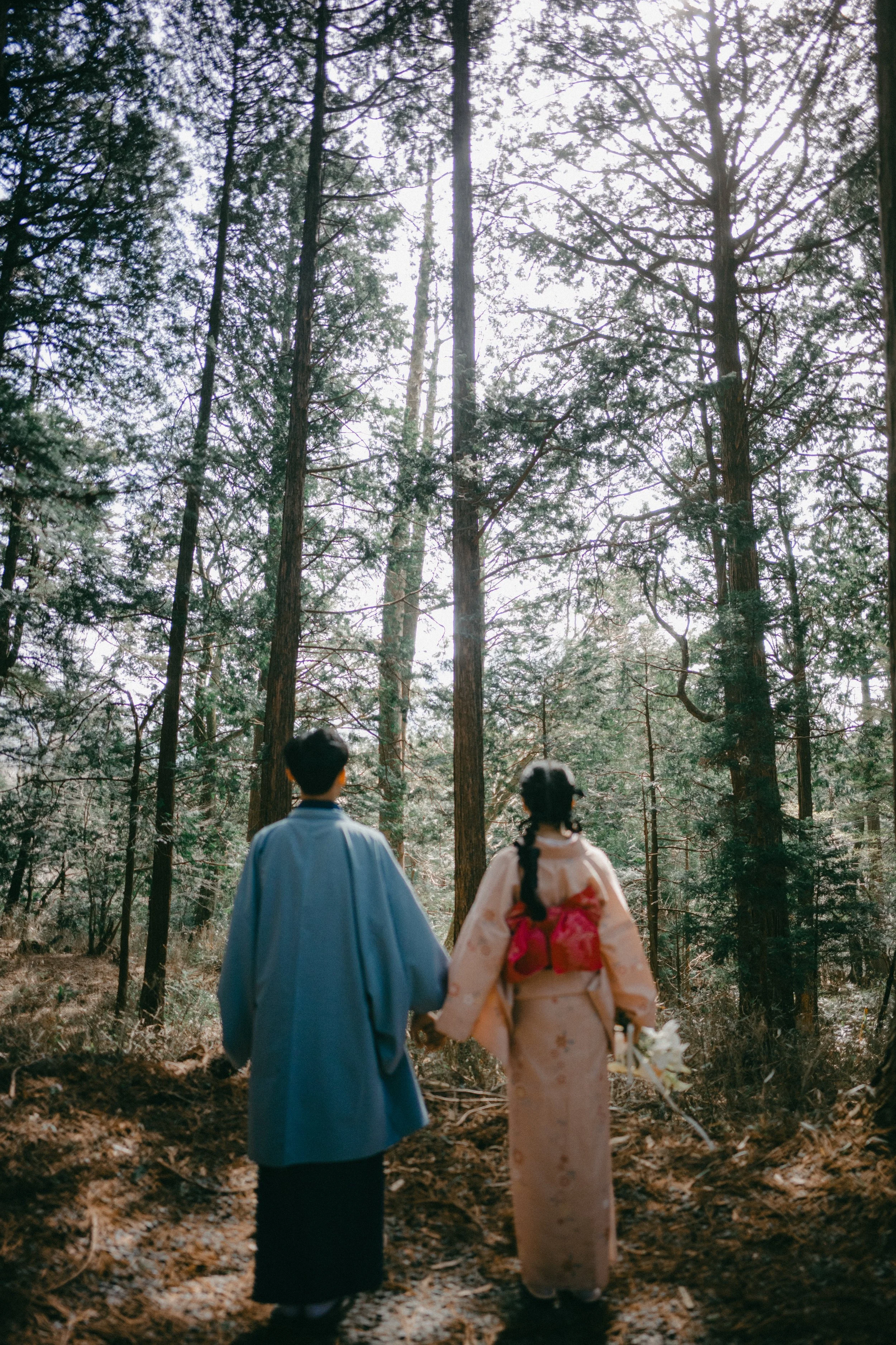A couple dressed in traditional Japanese wedding attire walking hand in hand through a forested area with tall trees and sunlight filtering through.