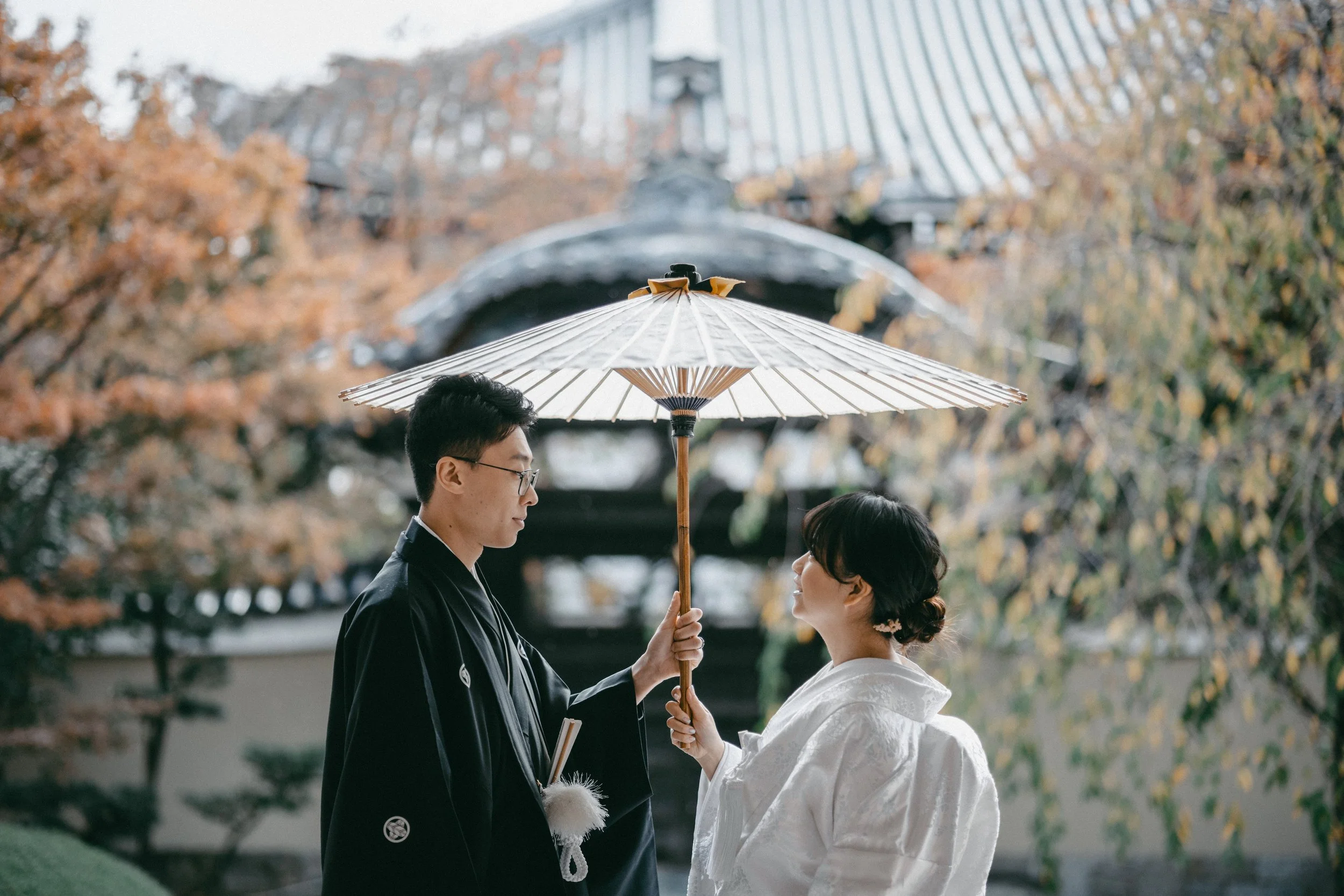 A couple dressed in traditional Japanese wedding attire stands outdoors, sharing a moment under a white parasol, with a garden and traditional building in the background.