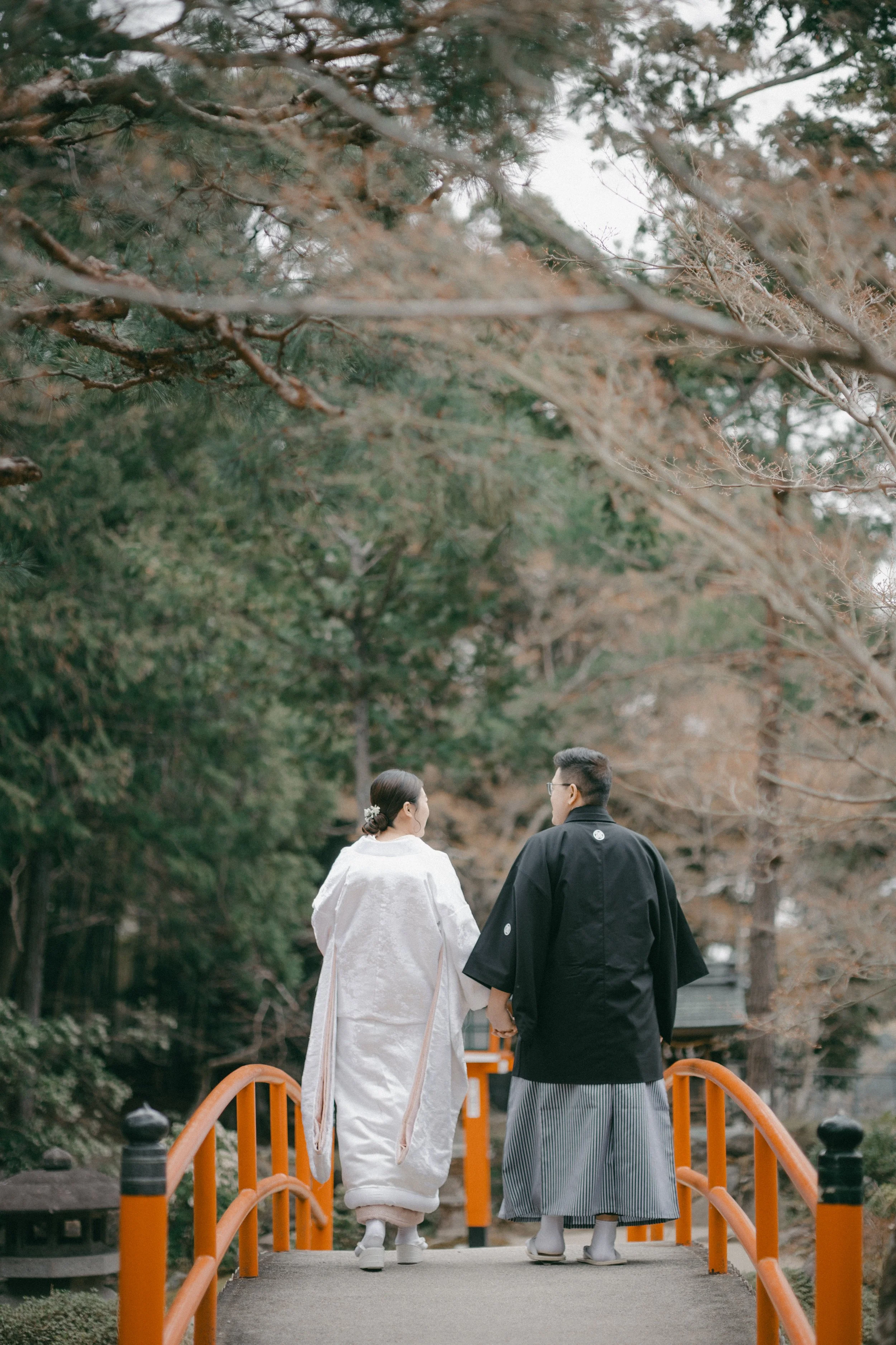 A couple walking on a small bridge in a wooded area, dressed in traditional Japanese wedding attire, surrounded by trees with autumn foliage.