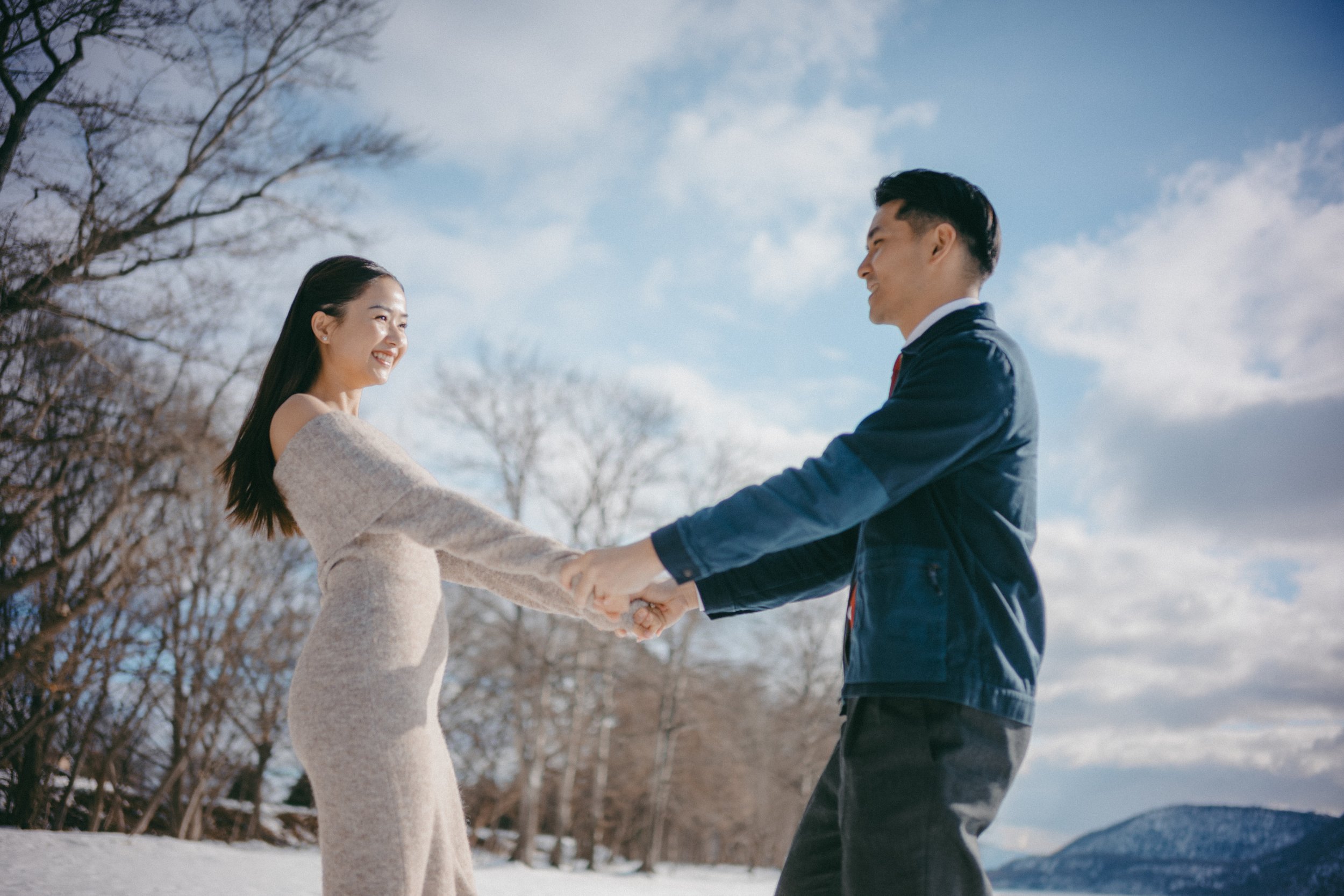 A couple holding hands and smiling outdoors in a snowy landscape with trees and a cloudy sky.