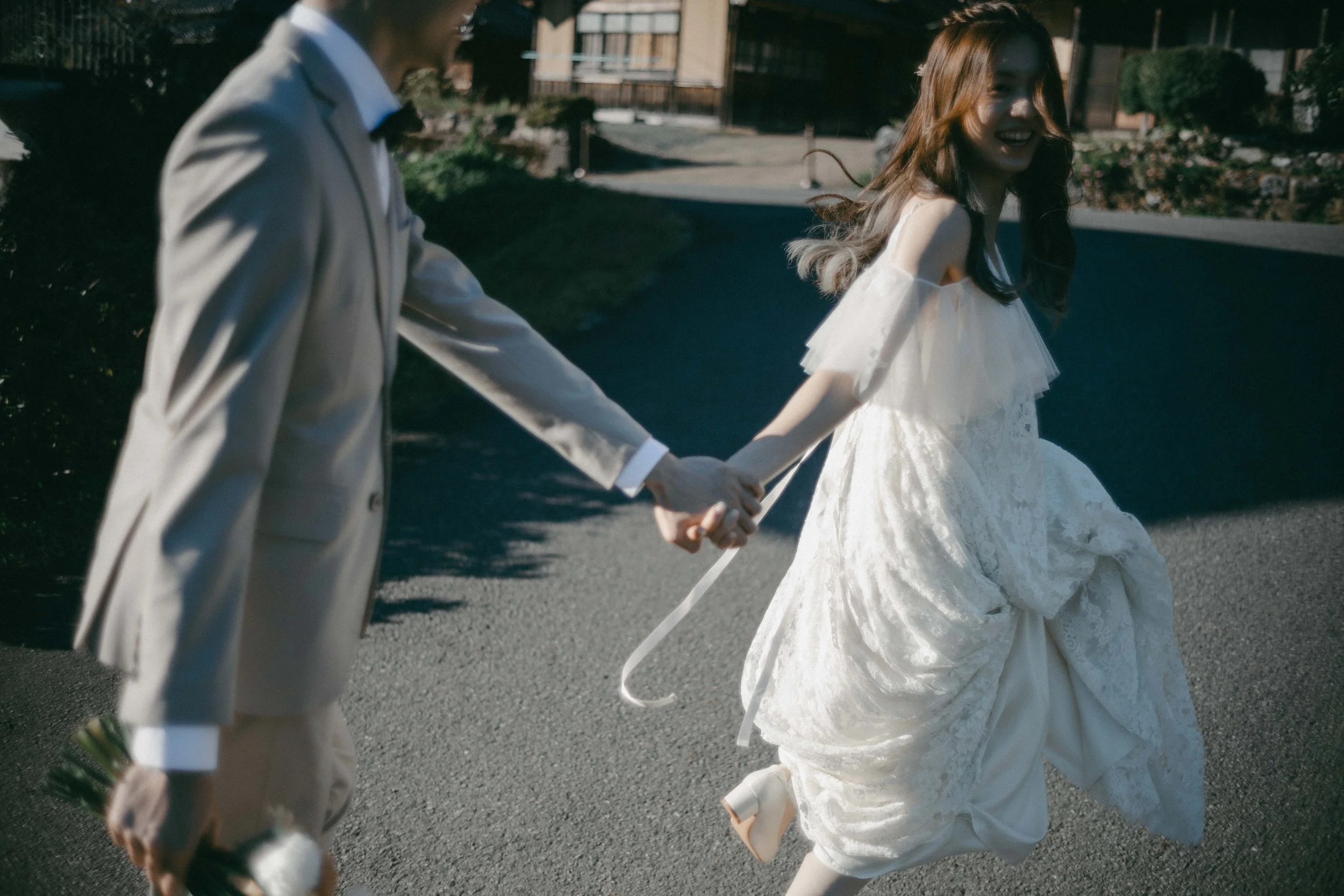A couple holding hands and running outdoors, with the woman wearing a white wedding dress and the man in a light-colored suit, during daytime.