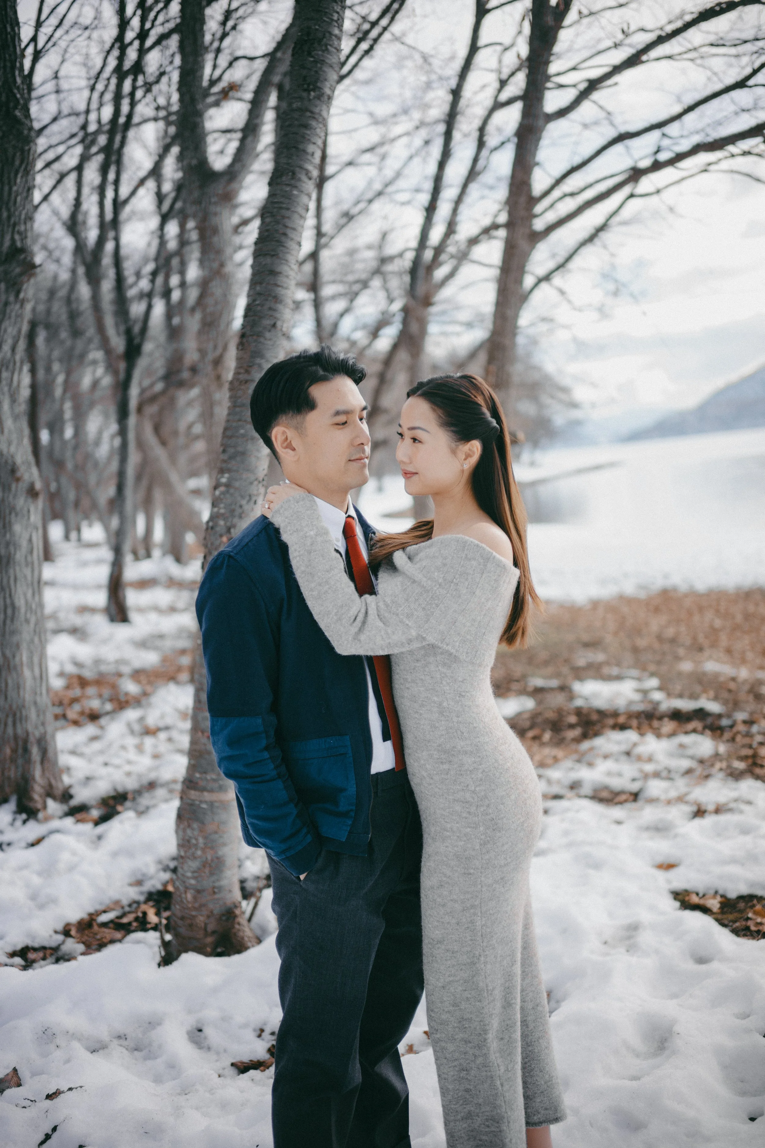A couple standing close together outdoors in a snowy forest, with leafless trees and a lake in the background. The woman is wearing a form-fitting gray dress, and the man is dressed in a dark blue jacket, white shirt, and red tie.