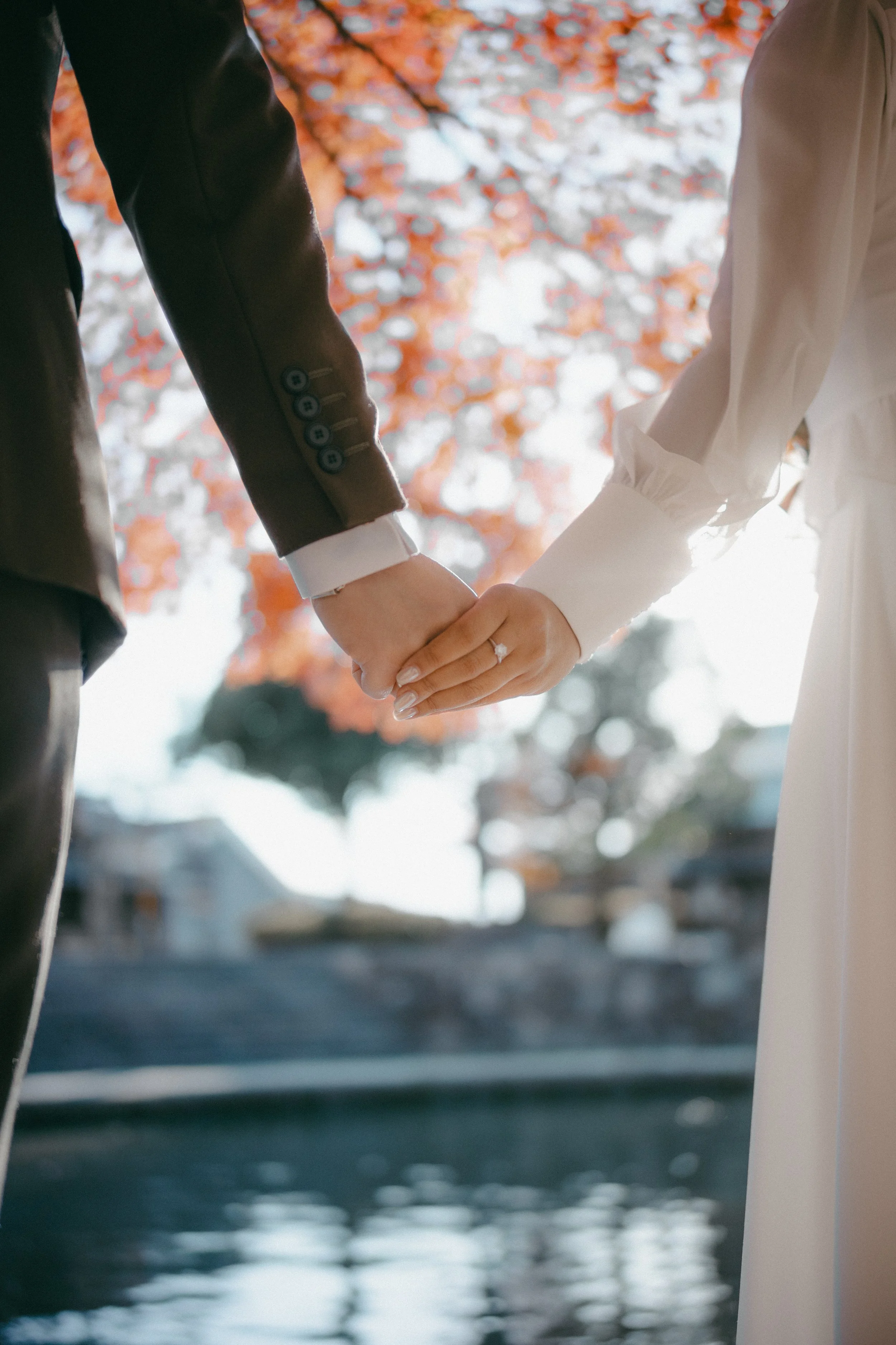 Close-up of a bride and groom holding hands outdoors with a blurred background of orange autumn leaves and water.